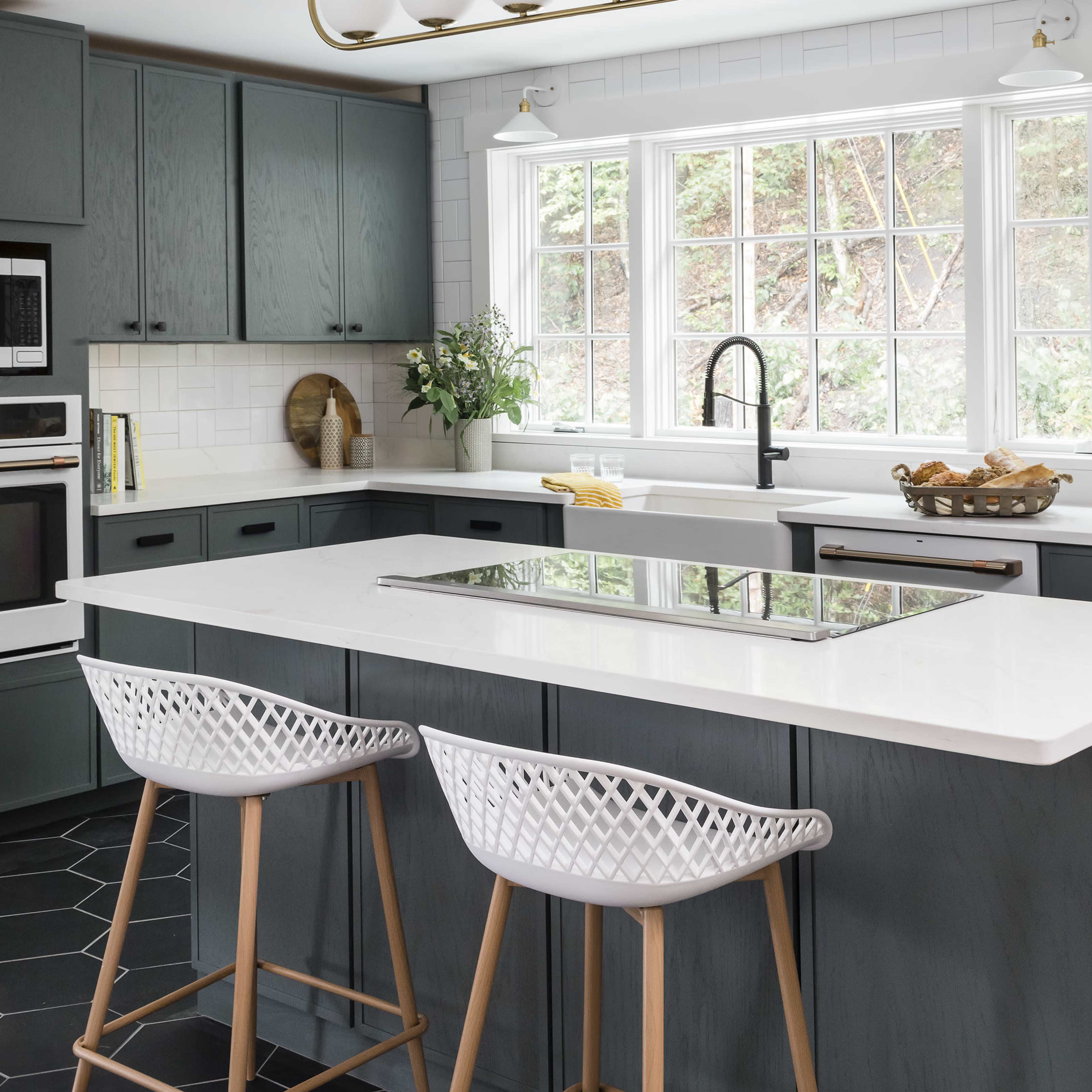 A modern kitchen features a large island with two wicker bar stools, under a stylish light fixture, and a window that provides natural light.