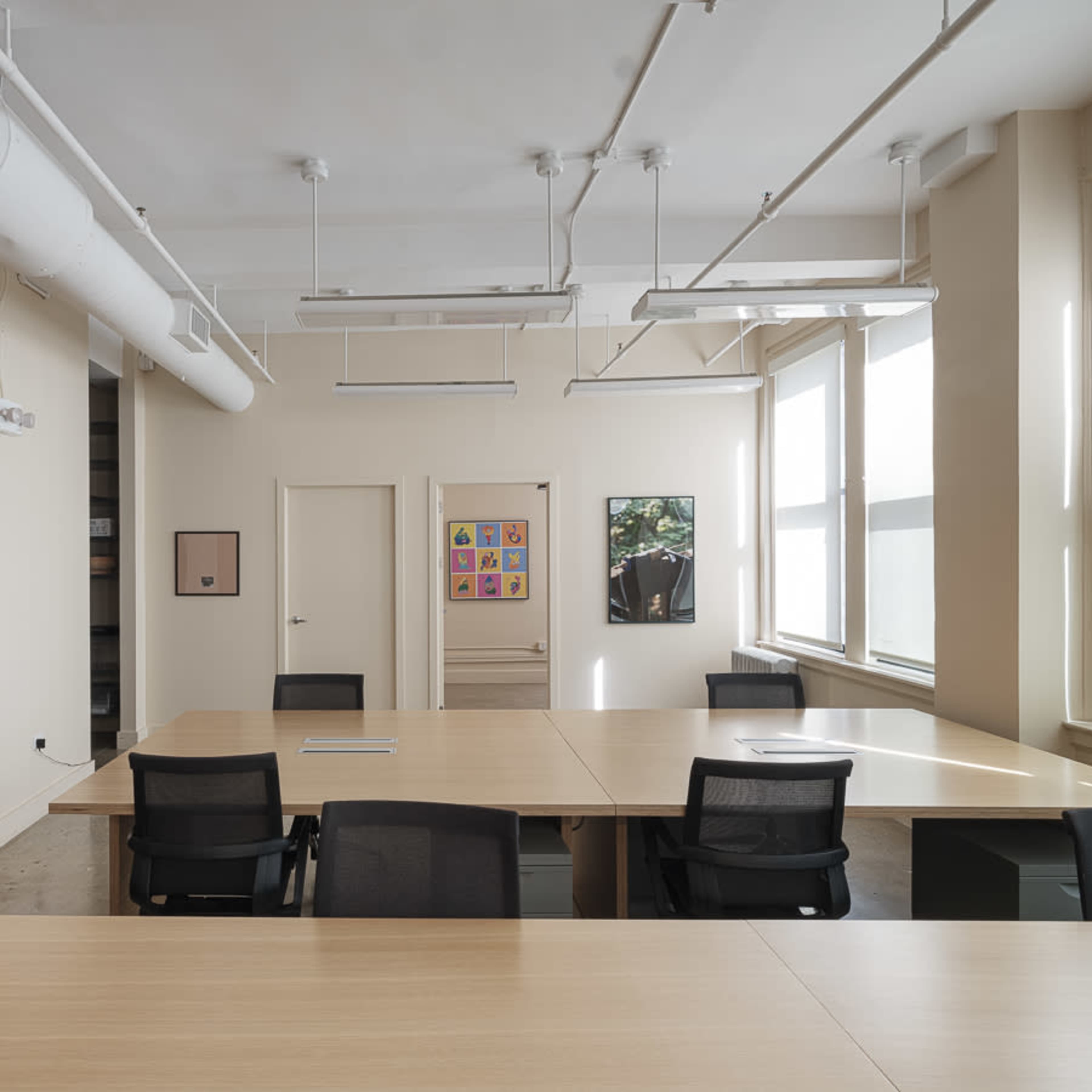 A modern office space featuring a large central table surrounded by black chairs, with art displayed on the walls and ample natural light from the windows.