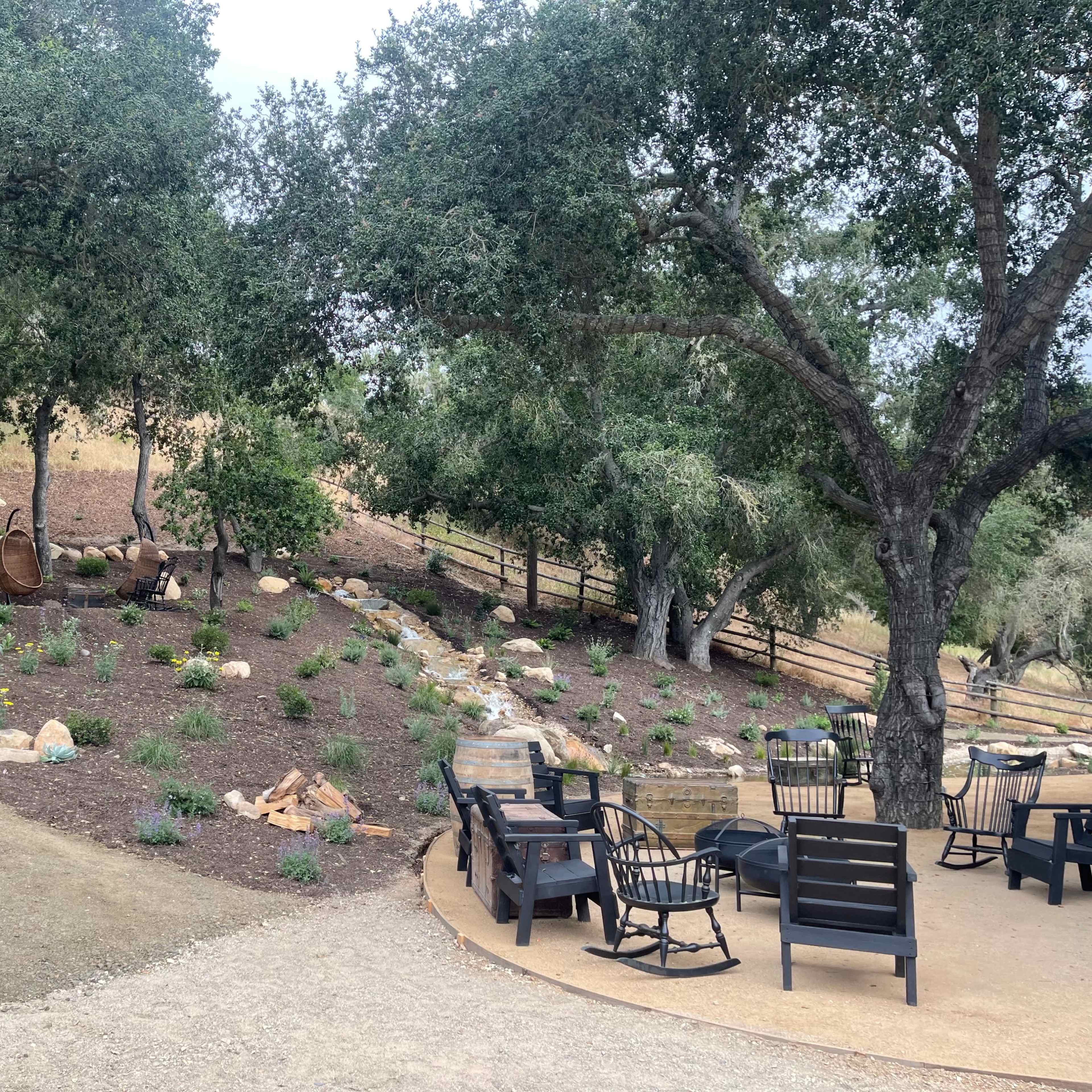 A landscaped area with a gravel path, seating arrangements, and trees, alongside a small stone water feature.