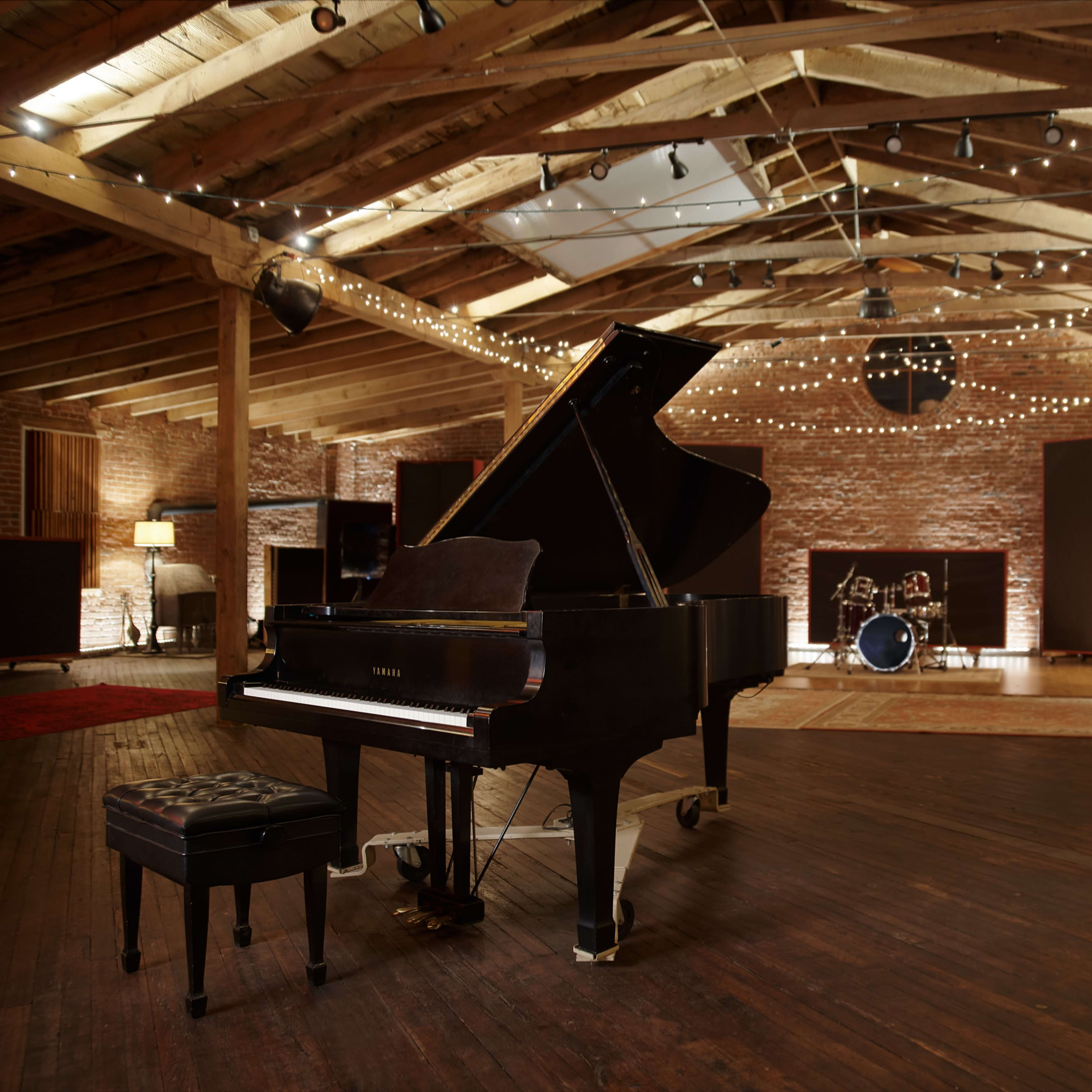 A grand piano is positioned on a wooden floor in a spacious, lit recording studio featuring exposed beams and a small drum set in the background.