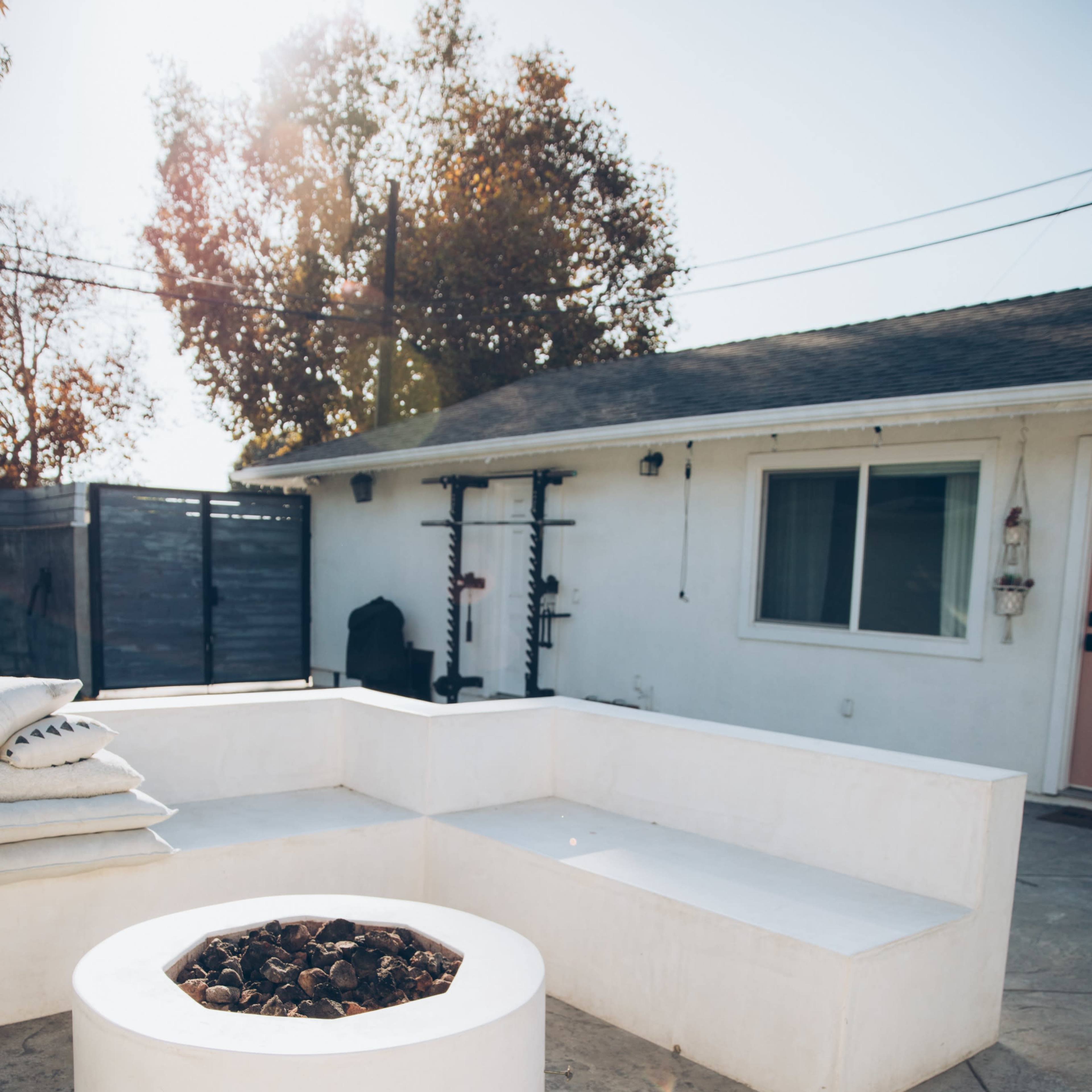 A backyard area with a circular fire pit surrounded by a white, built-in seating arrangement and a house in the background.