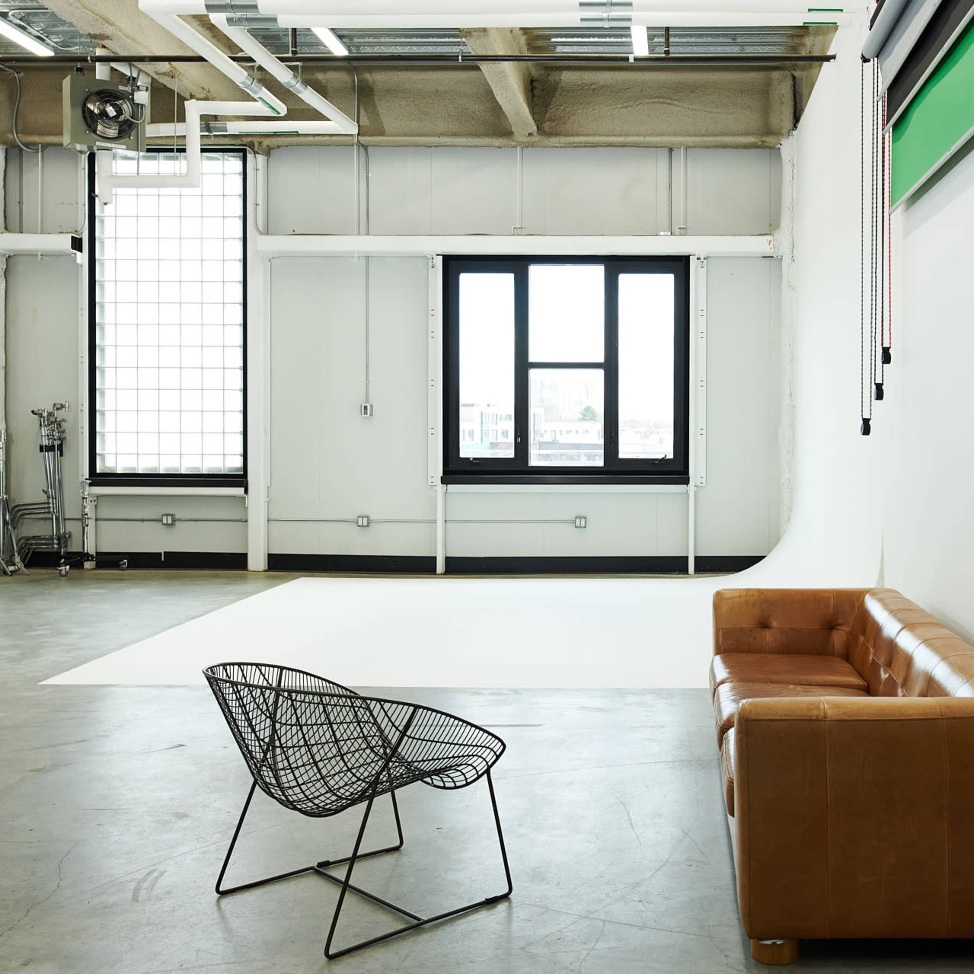A spacious photography studio with a white backdrop, a large window, and a brown leather couch paired with a wire chair.
