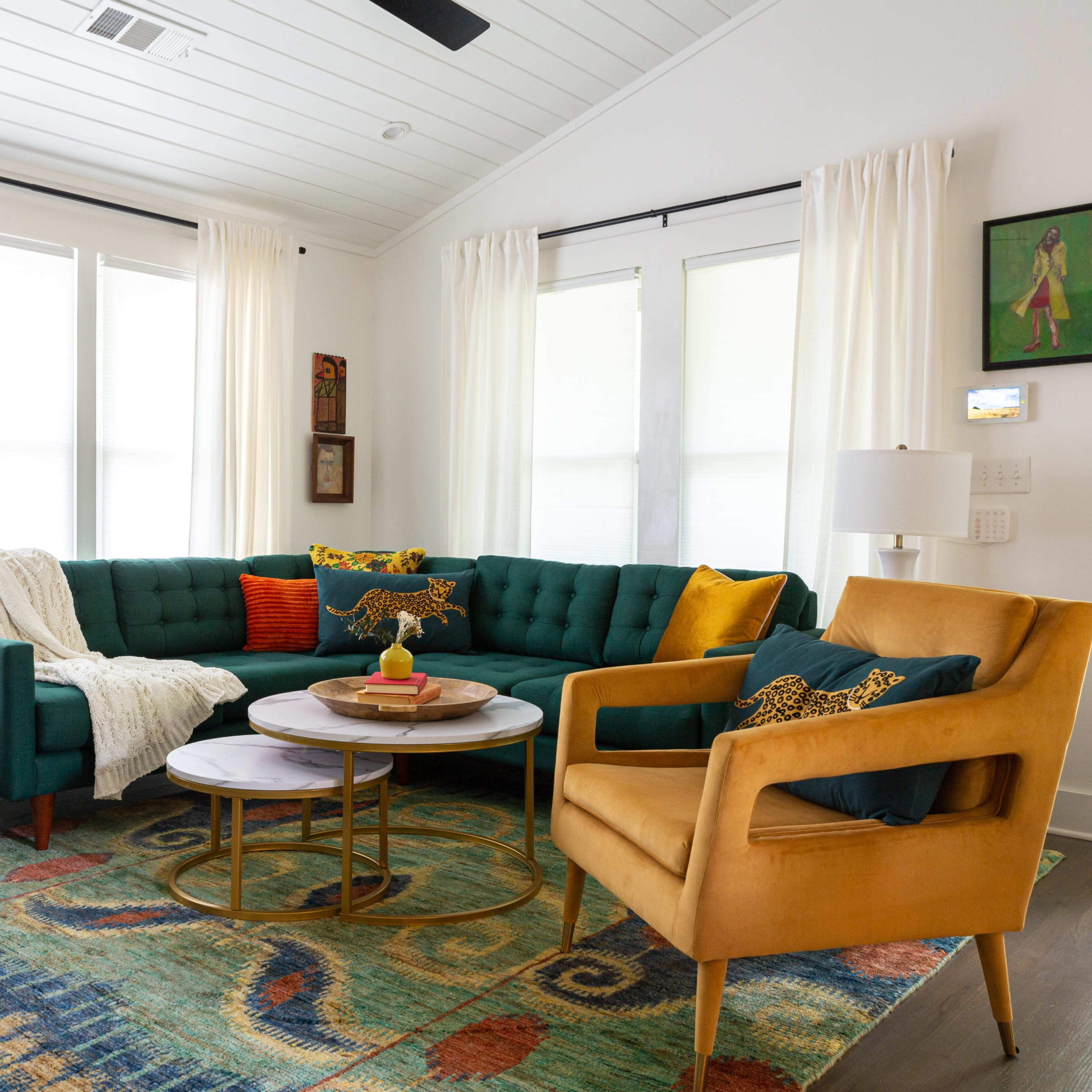 The living room features a green sectional couch, two lamps, a round coffee table, and a colorful area rug under bright windows.