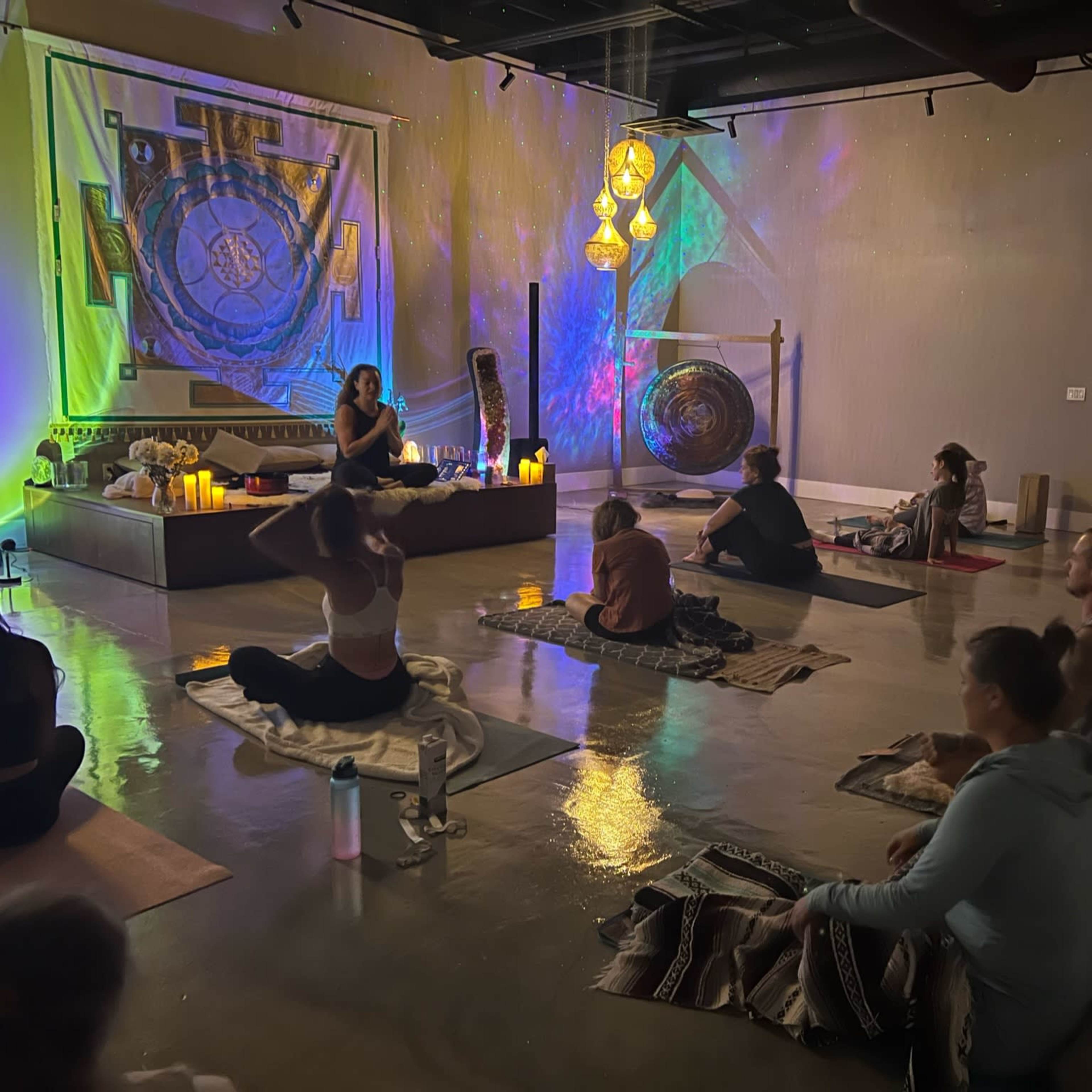 A group of individuals sits on yoga mats in a dimly lit studio, while a person leading the session is positioned at the front near candles and a large gong.