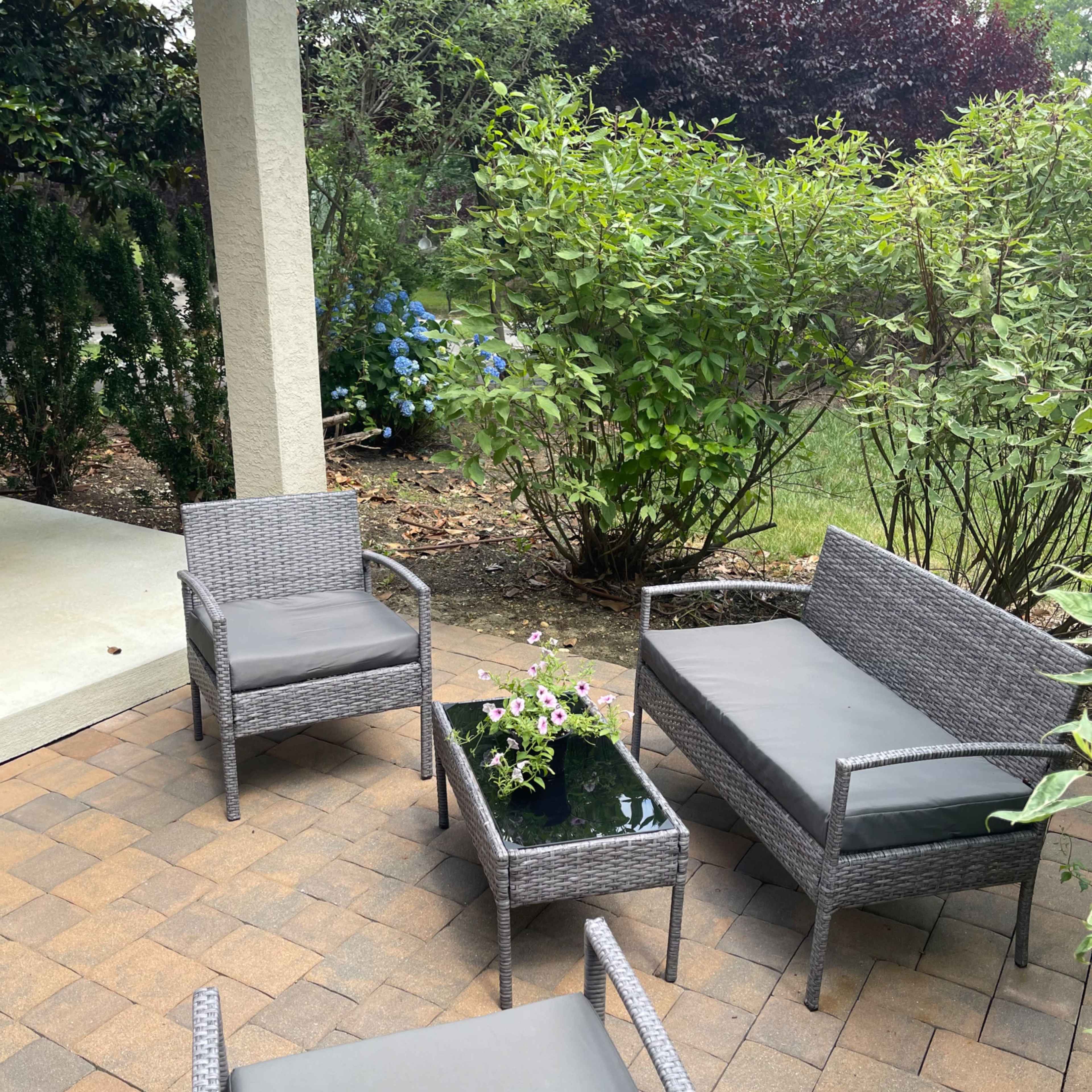 A set of gray outdoor furniture, consisting of two chairs, a loveseat, and a coffee table, is arranged on a stone patio surrounded by greenery.