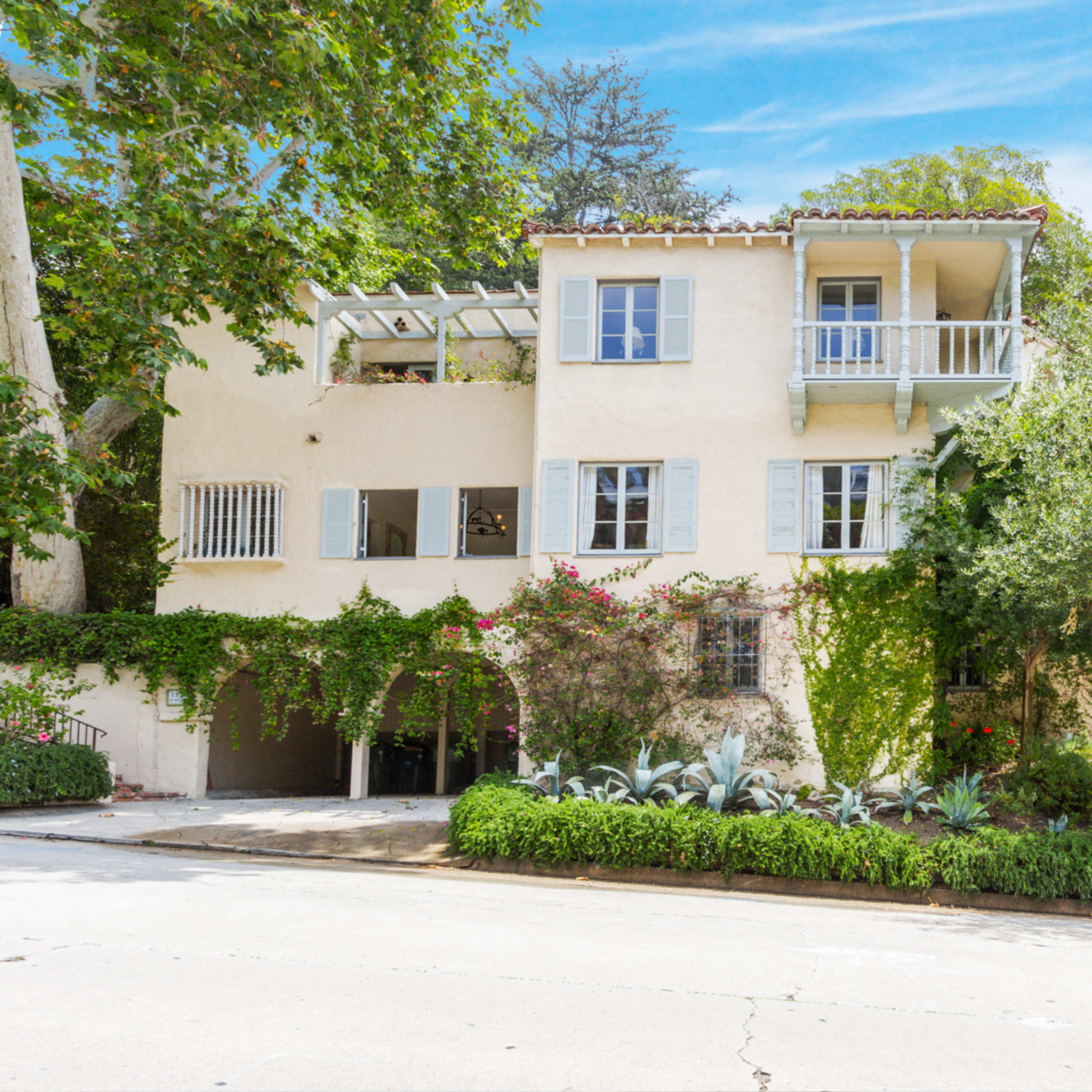 A two-story house with a Mediterranean design features balconies, greenery climbing its walls, and is surrounded by landscaped gardens on a quiet street.