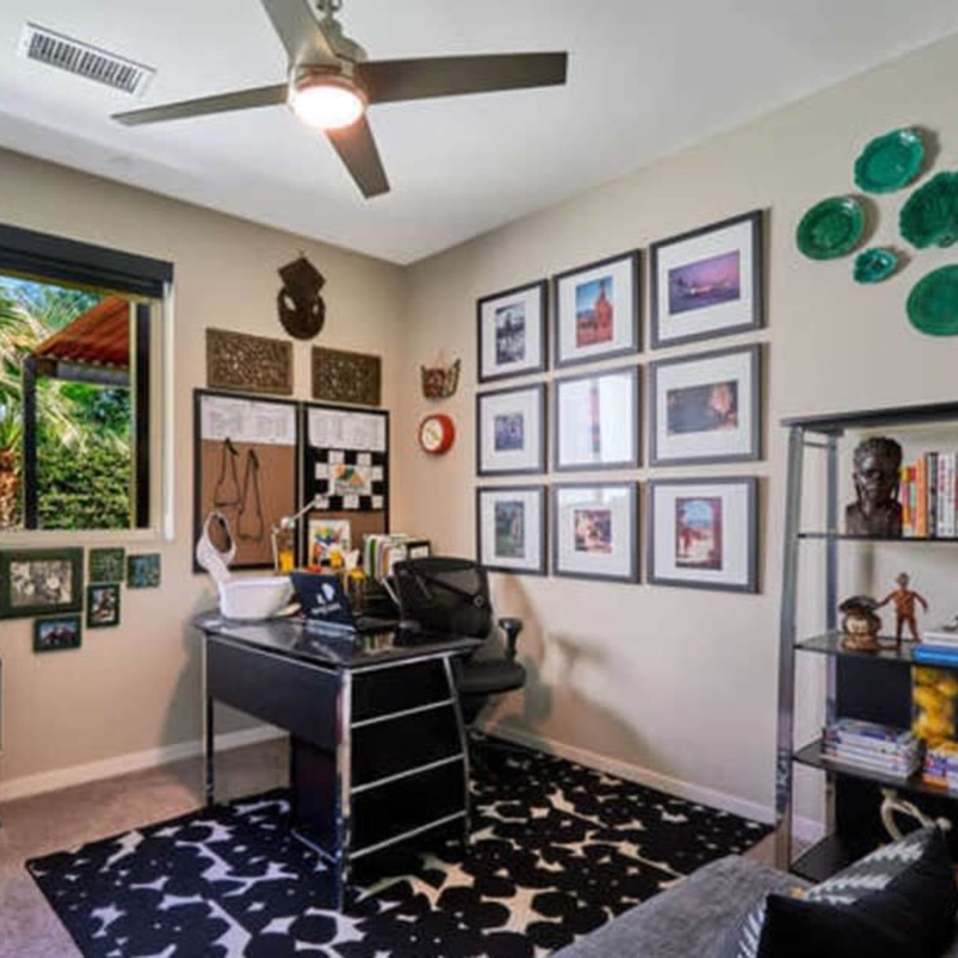 A home office with a black desk, a bookshelf, framed photos on the walls, and large windows overlooking a garden.