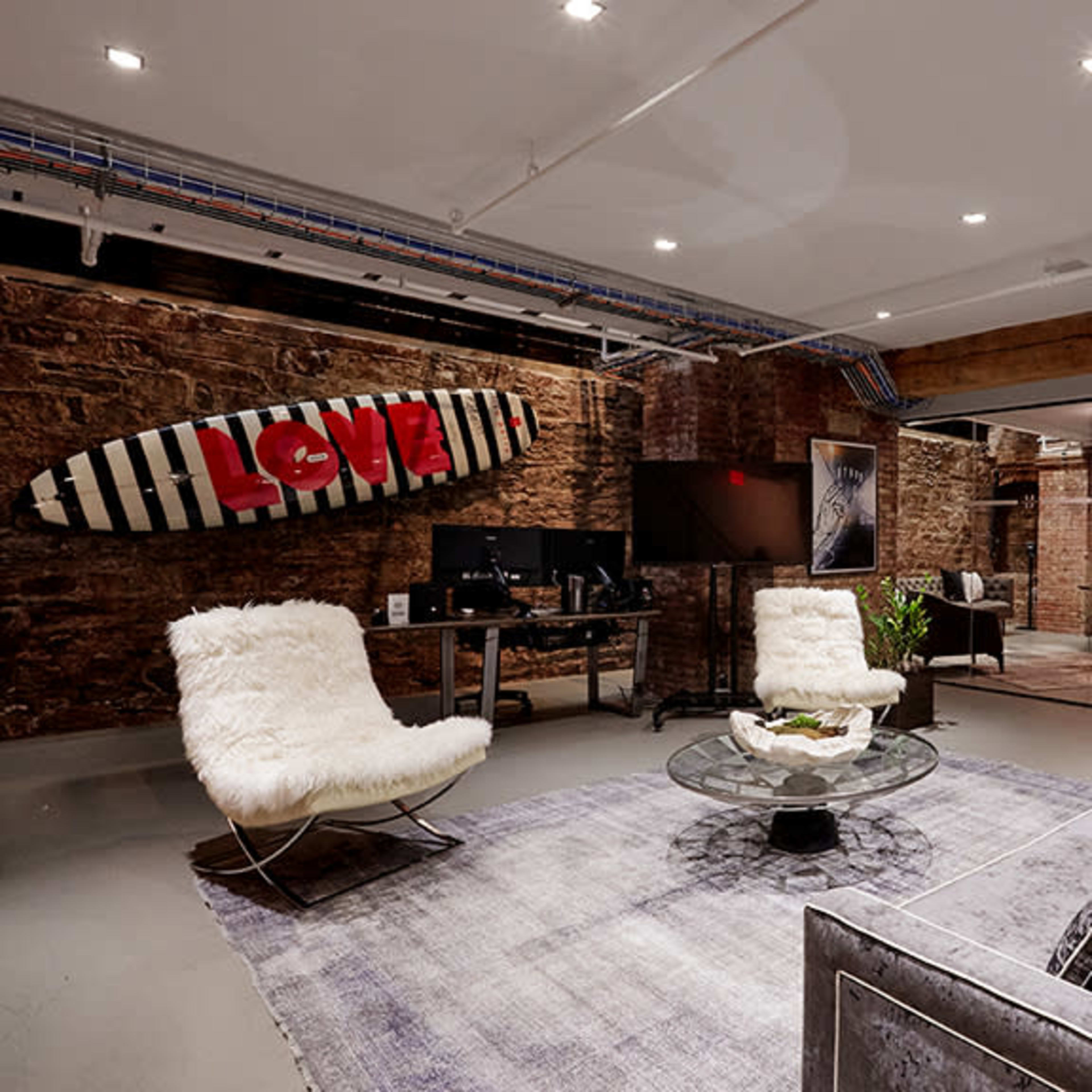 A modern industrial-style living space featuring a glass coffee table on a rug, two fur-covered chairs, a gray sofa, and a decorative surfboard on the wall.