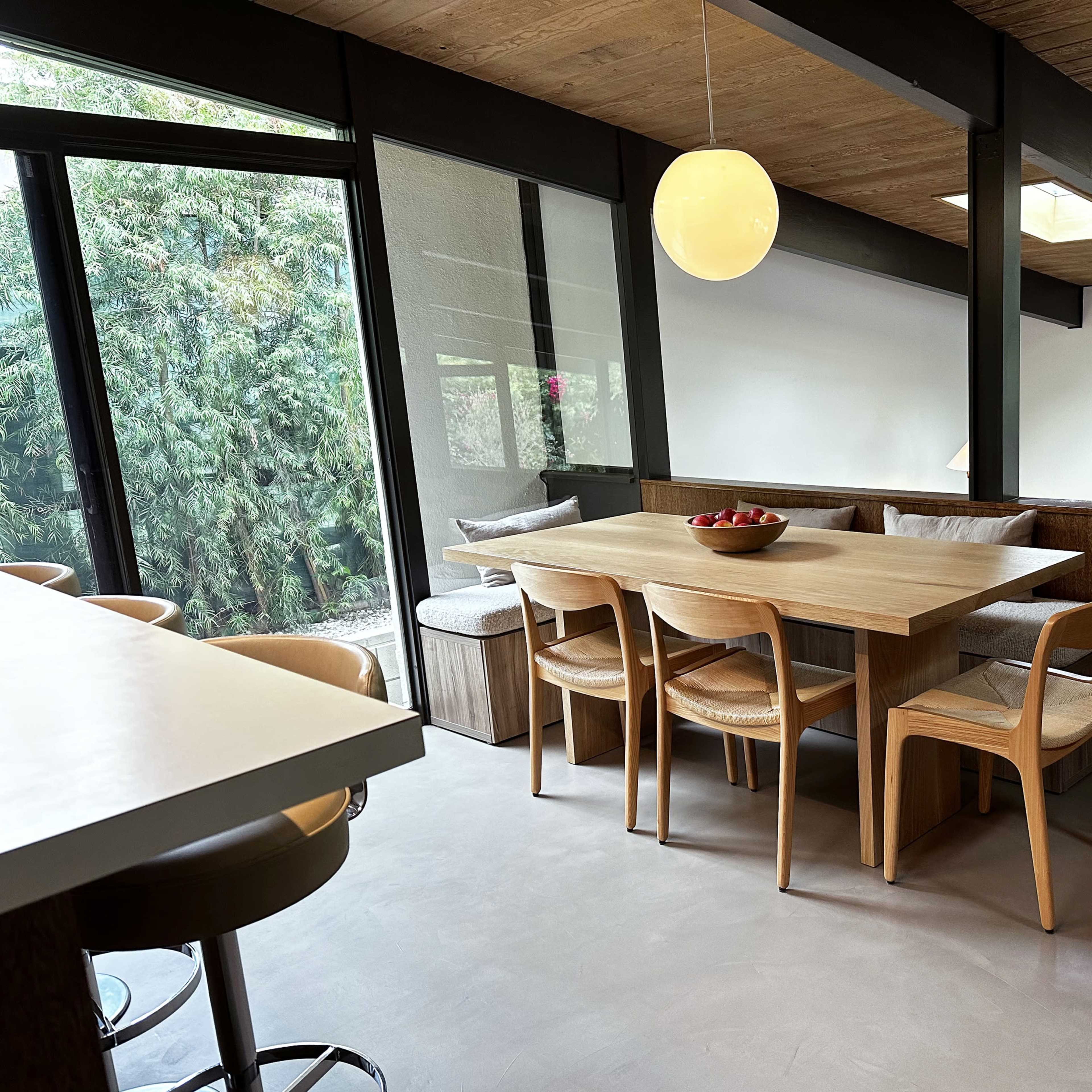 A modern dining area features a wooden table surrounded by chairs, with large windows providing views of greenery outside.