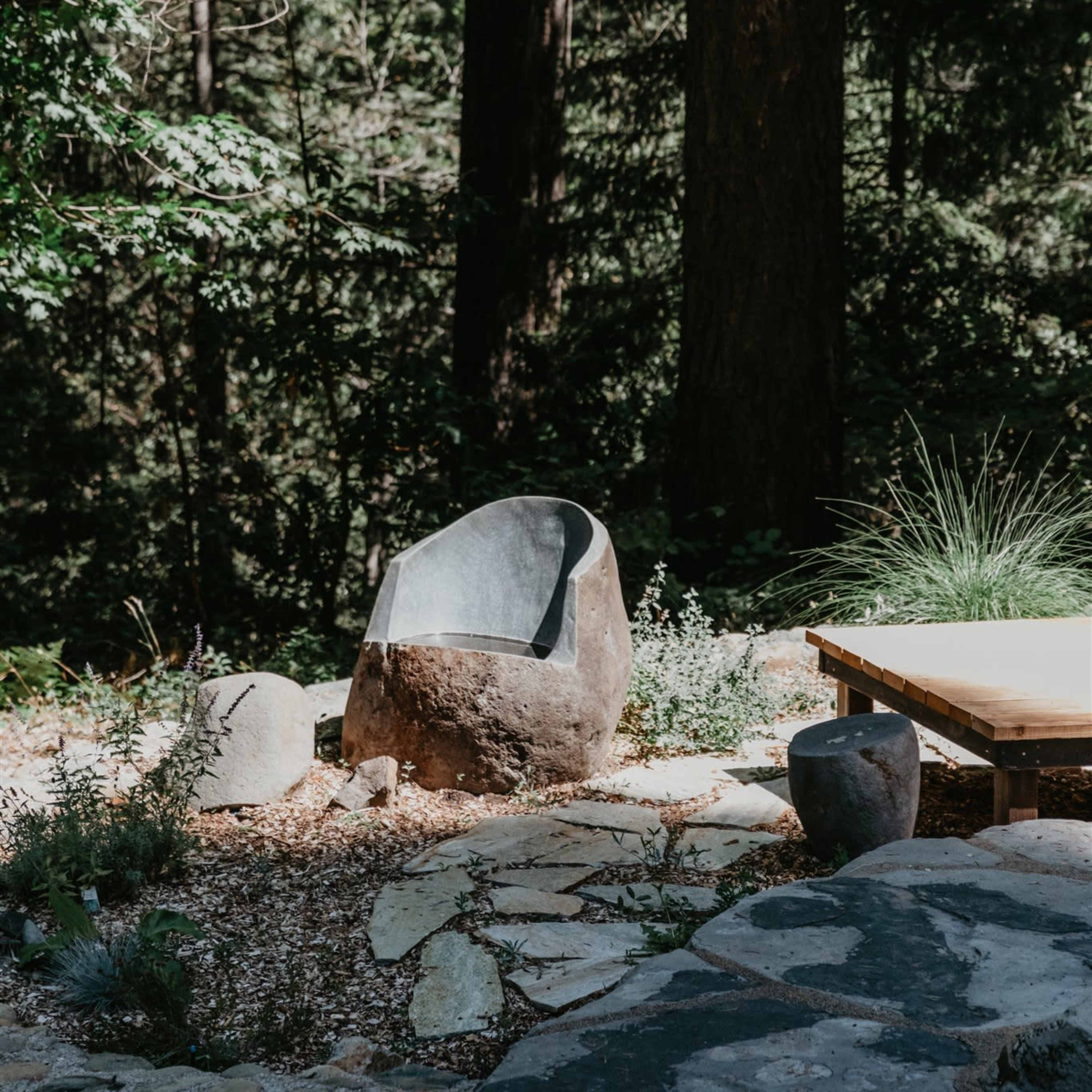 A large stone chair and several smaller stones are situated on a stone pathway in a forested area.