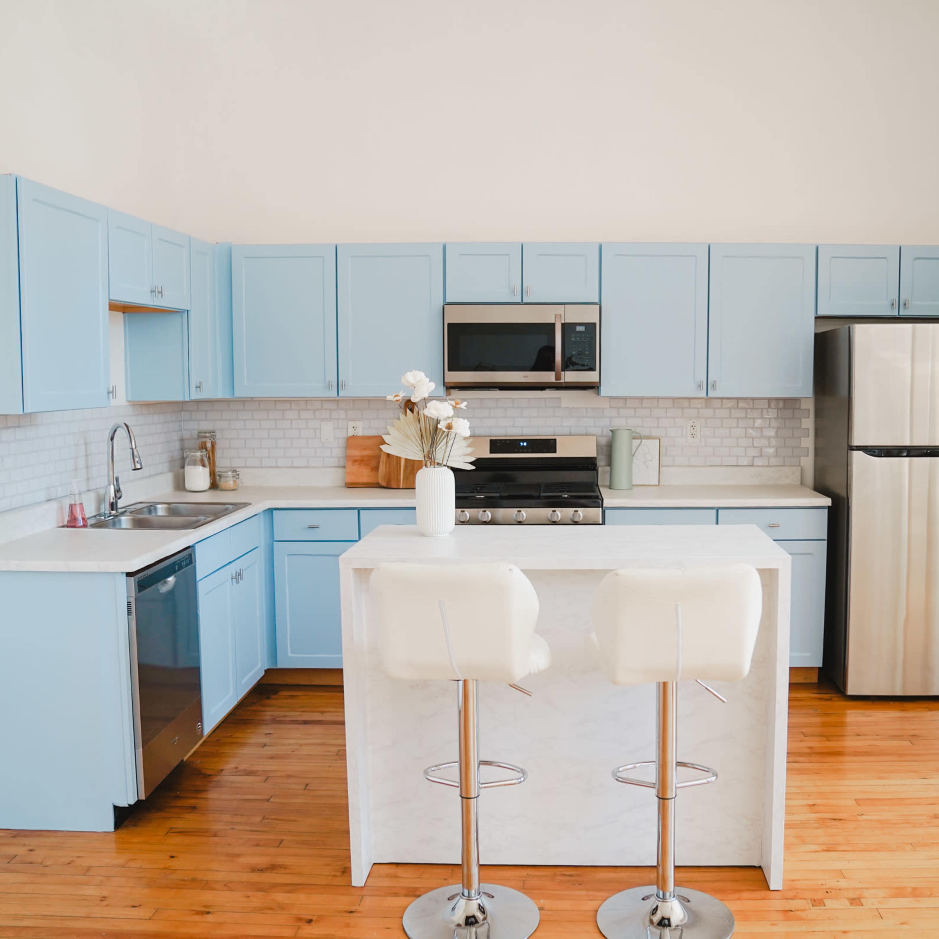 A modern kitchen features light blue cabinetry, stainless steel appliances, and a central island with two bar stools.