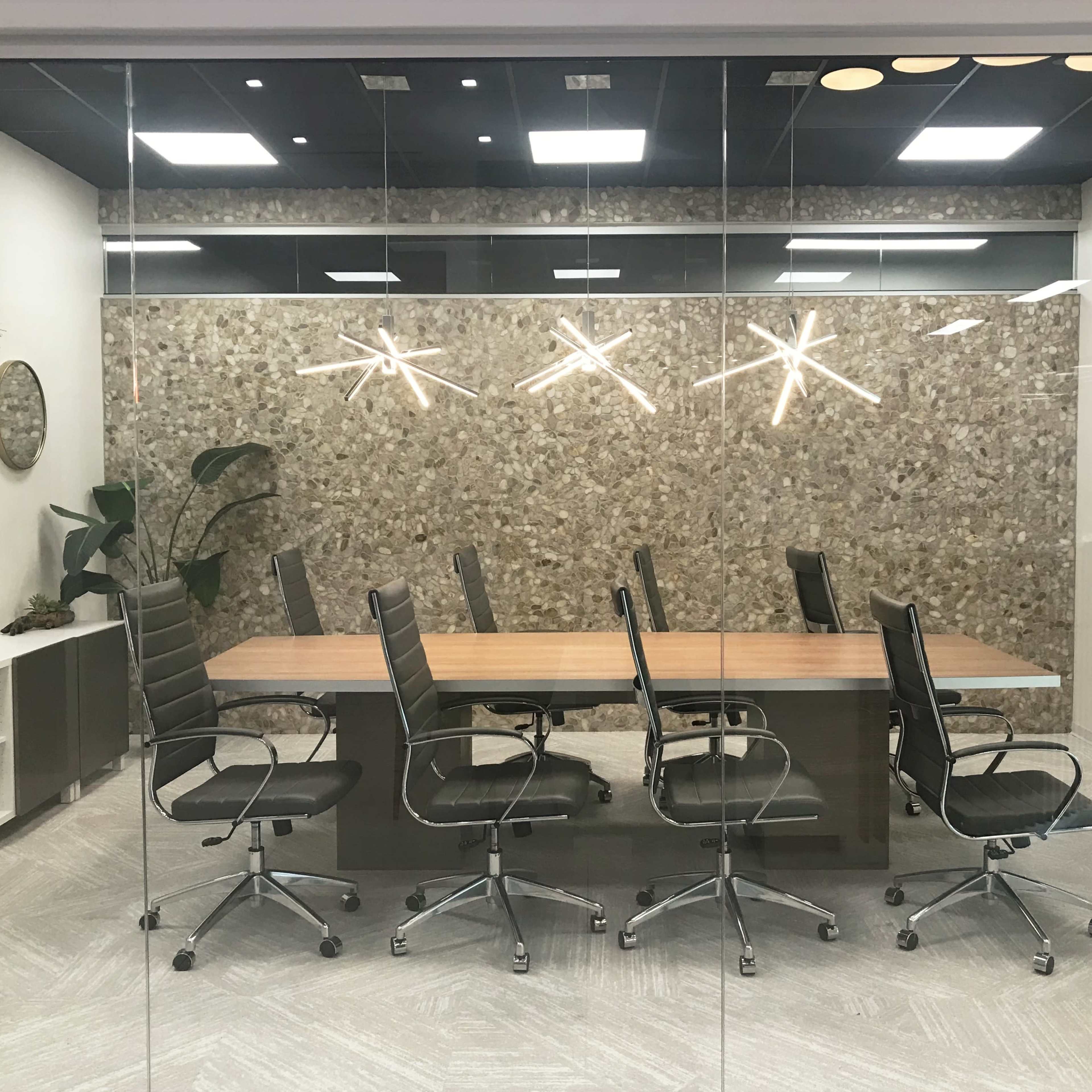 A modern conference room with a long wooden table surrounded by six black executive chairs, enclosed by a glass wall.