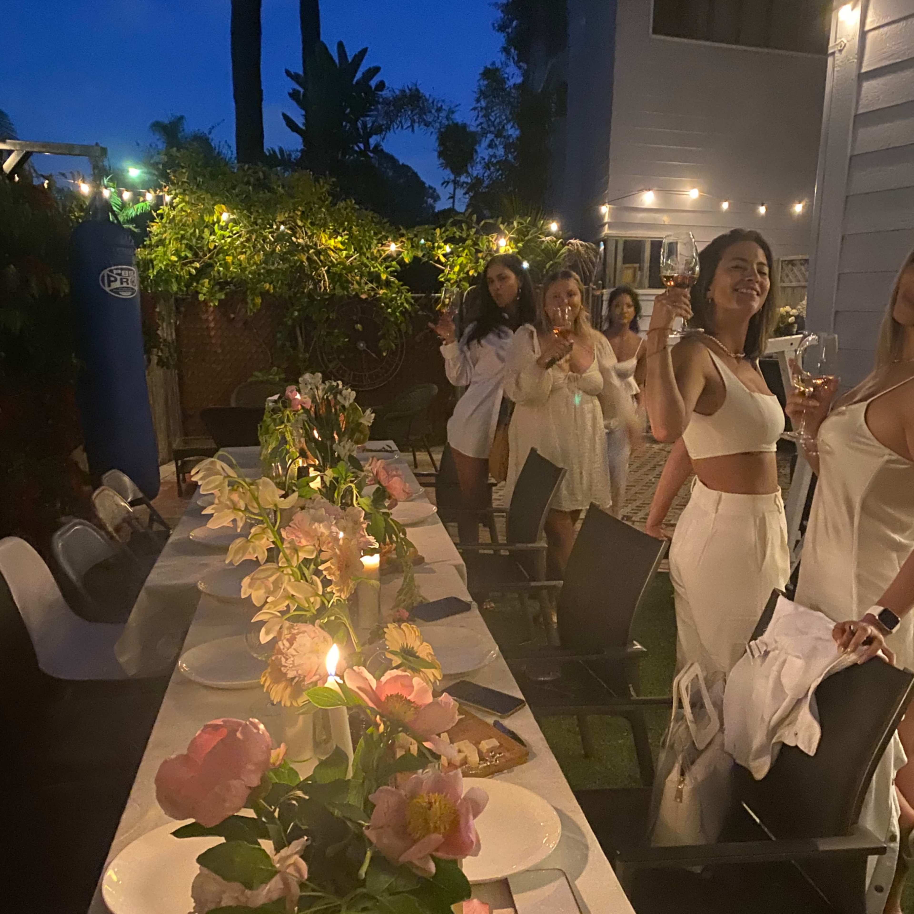 A group of people in white attire raises glasses in a garden setting during a nighttime dinner party, with a long table adorned with flowers and candles.