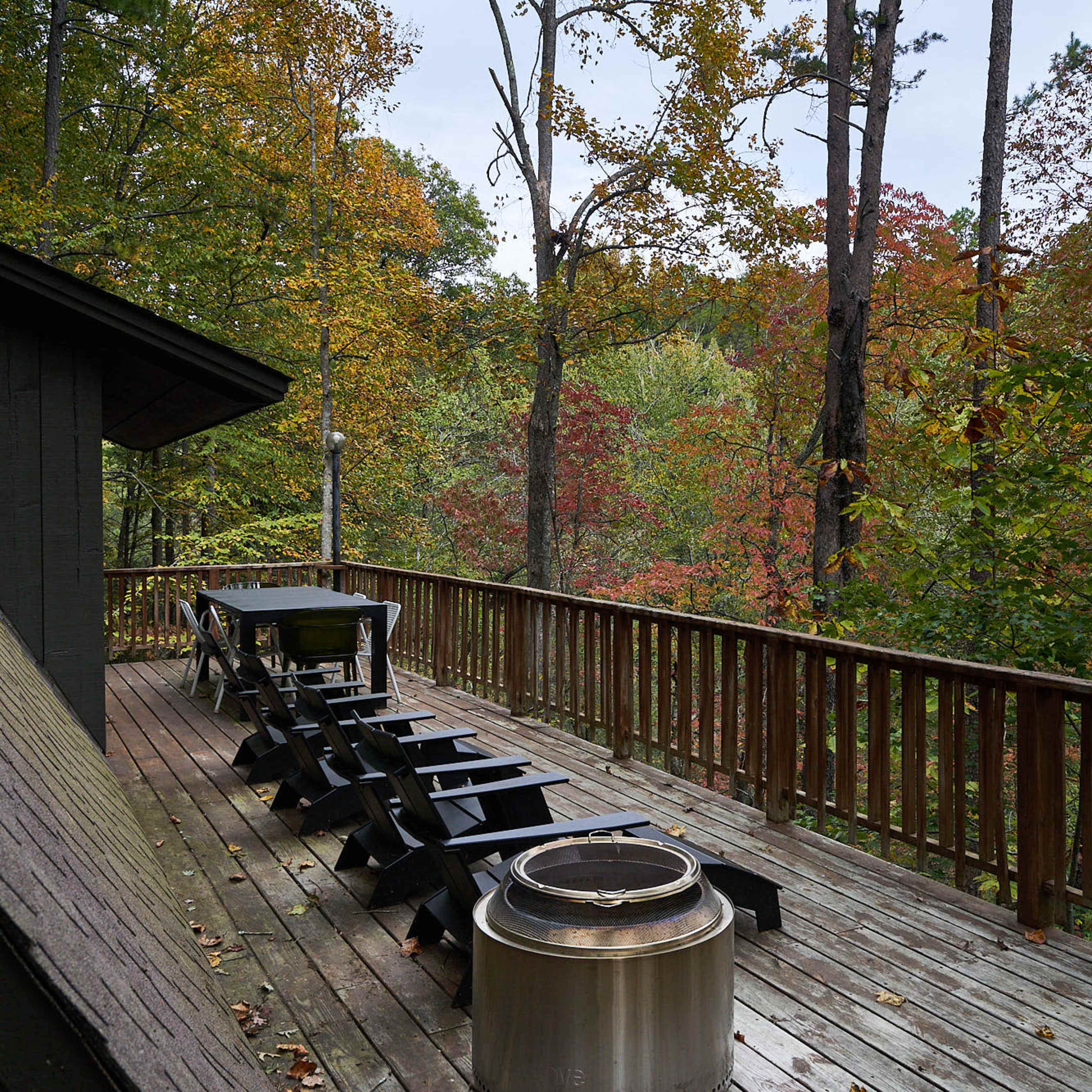A wooden deck with a set of black chairs overlooks a forest of trees displaying autumn colors.