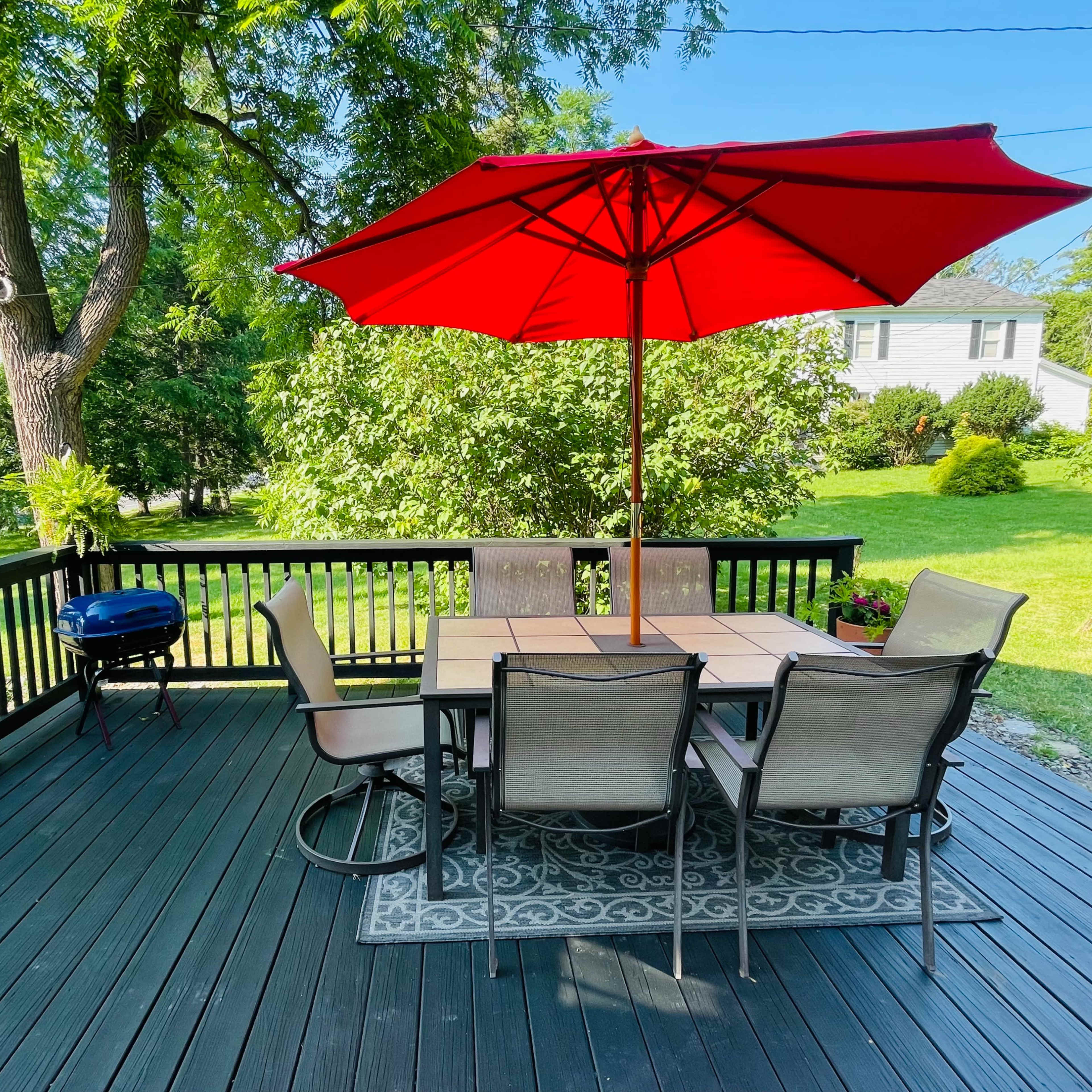 A patio with a round table and six chairs is topped by a red umbrella, overlooking a green garden with a barbecue grill nearby.