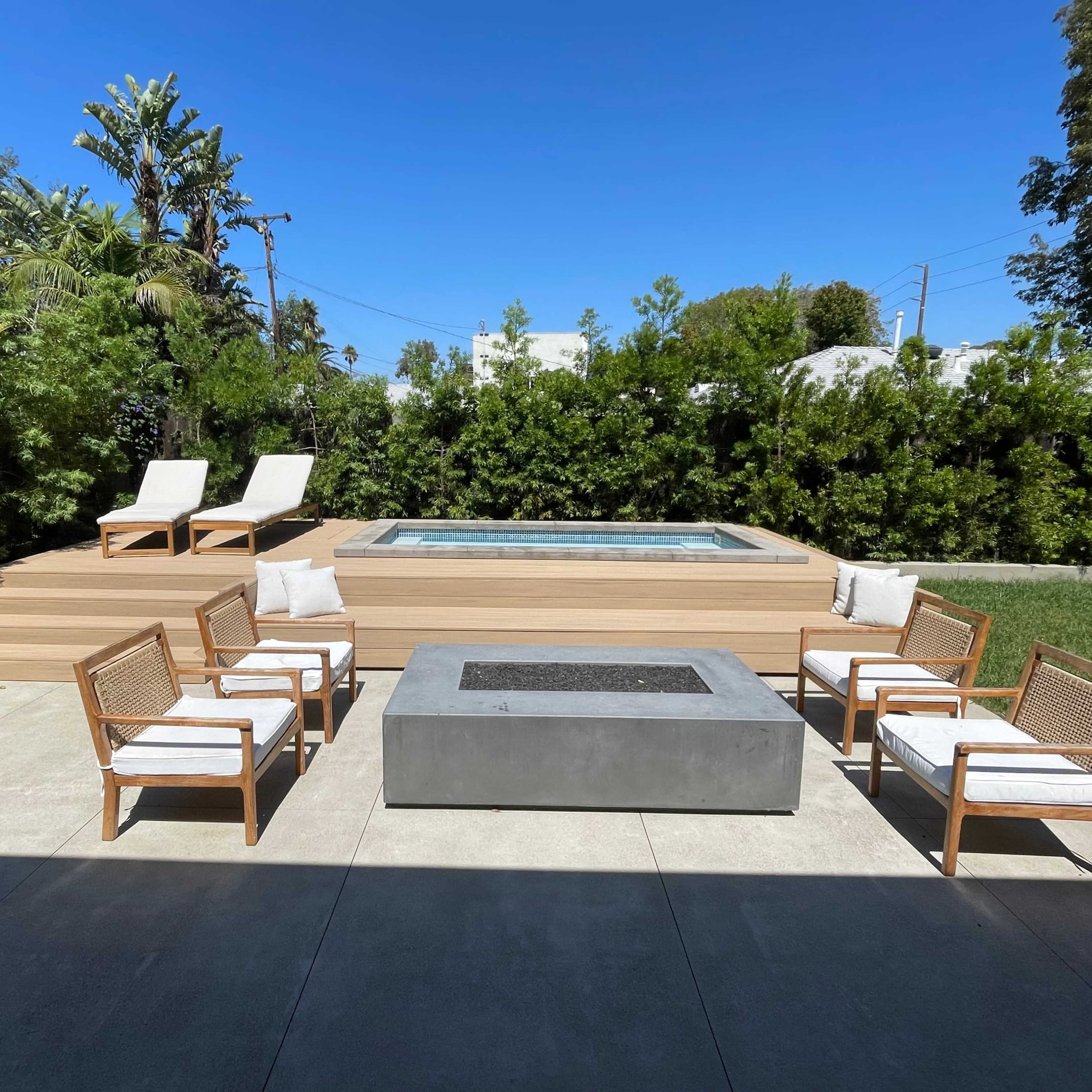 A landscaped backyard featuring a hot tub on a raised wooden platform, surrounded by seating areas and green foliage.