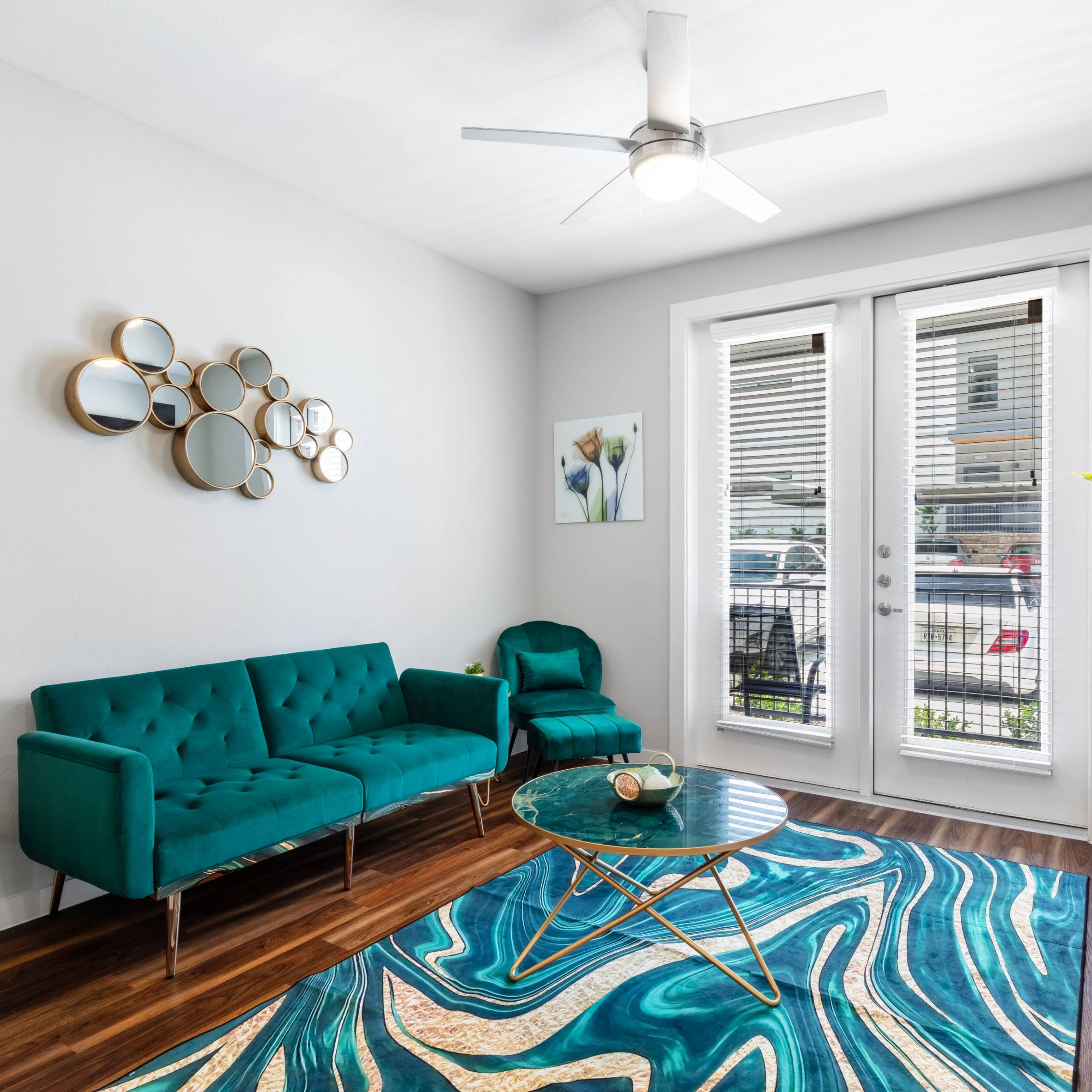A modern living room with a teal sofa, a glass coffee table on a patterned rug, and large windows opening to a balcony.