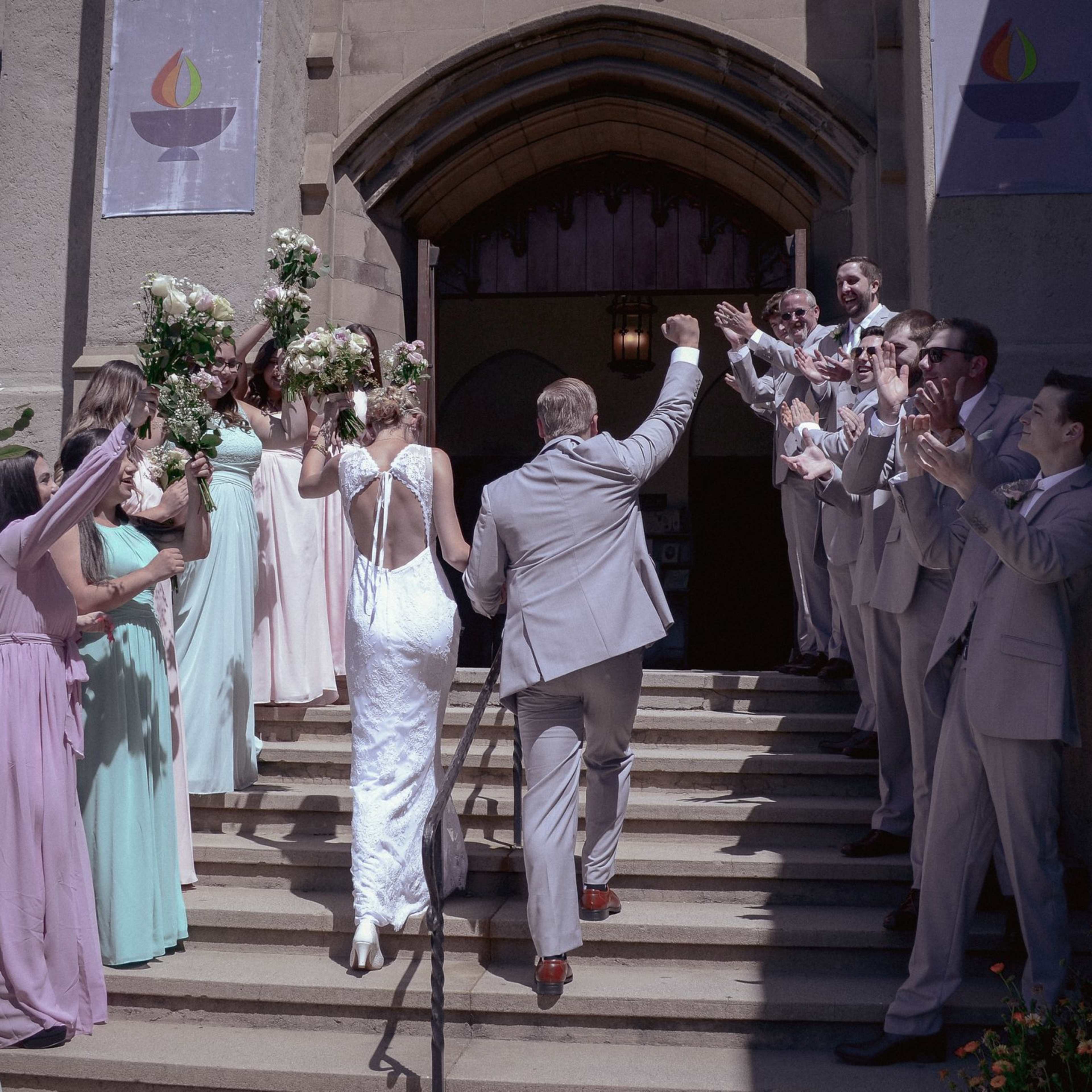 A wedding party stands on the steps of a church, with the bride and groom ascending while guests cheer and hold bouquets.