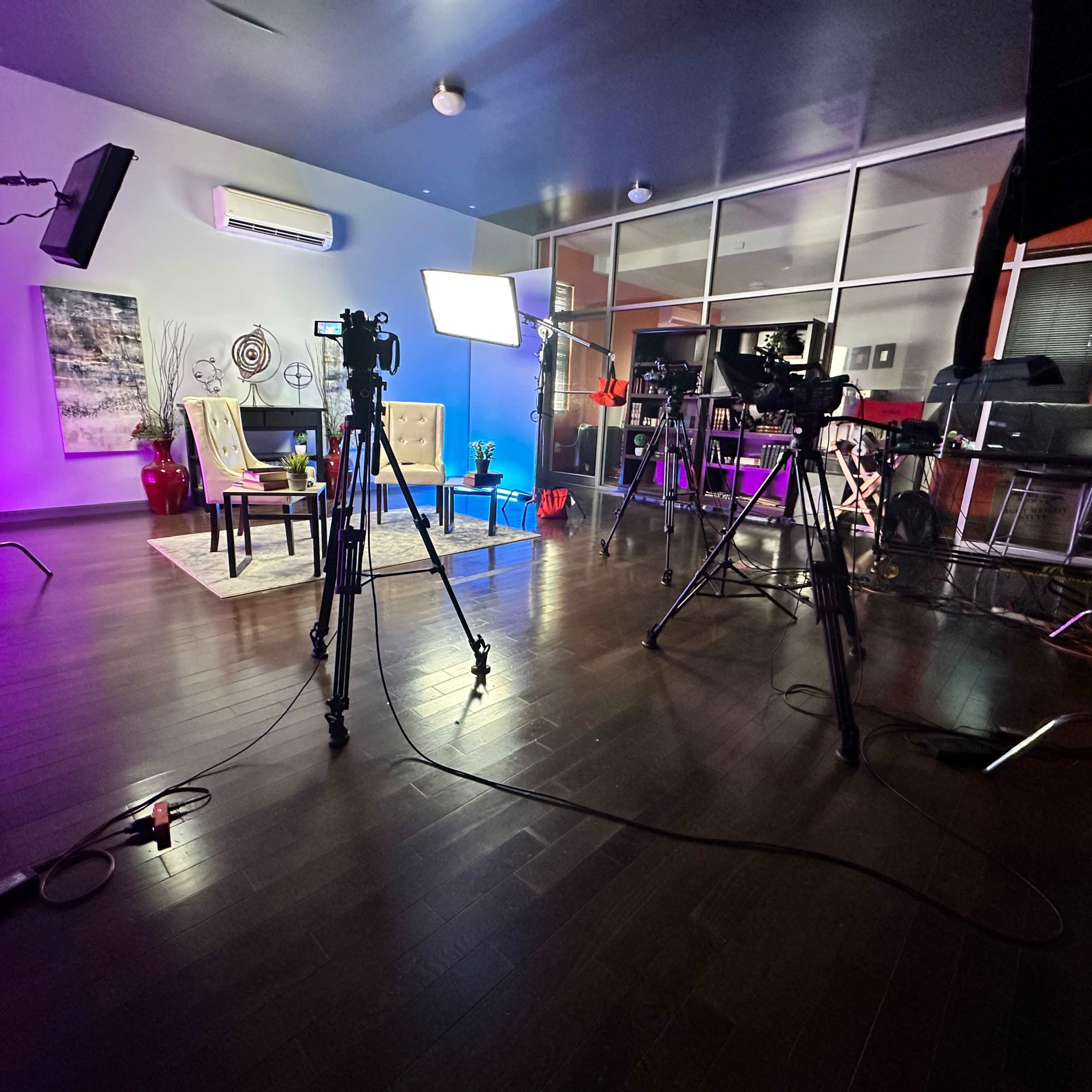 A well-lit studio setup with three cameras positioned on tripods facing a pair of chairs in front of a decorated backdrop.