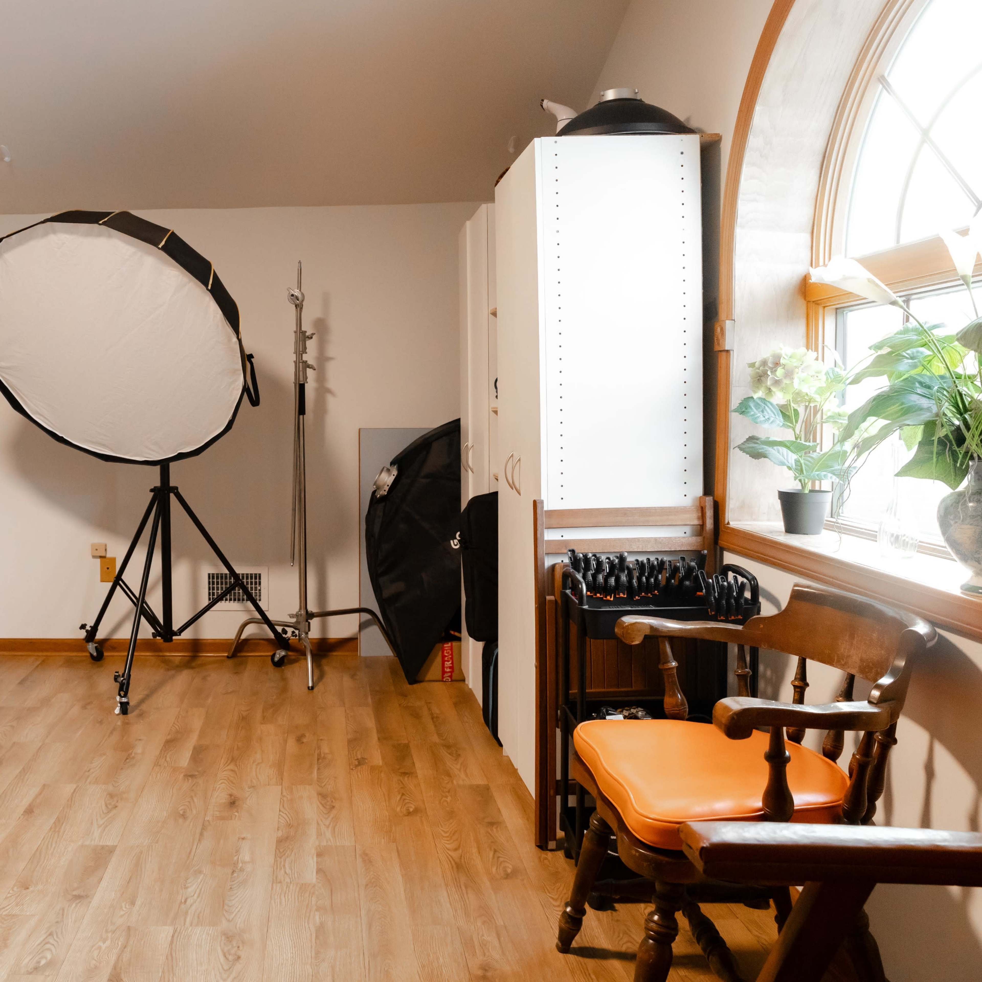A spacious room with wooden flooring, a large softbox light on a stand, a black chair, a monitor on a stand, shelving, and a potted plant near a window.