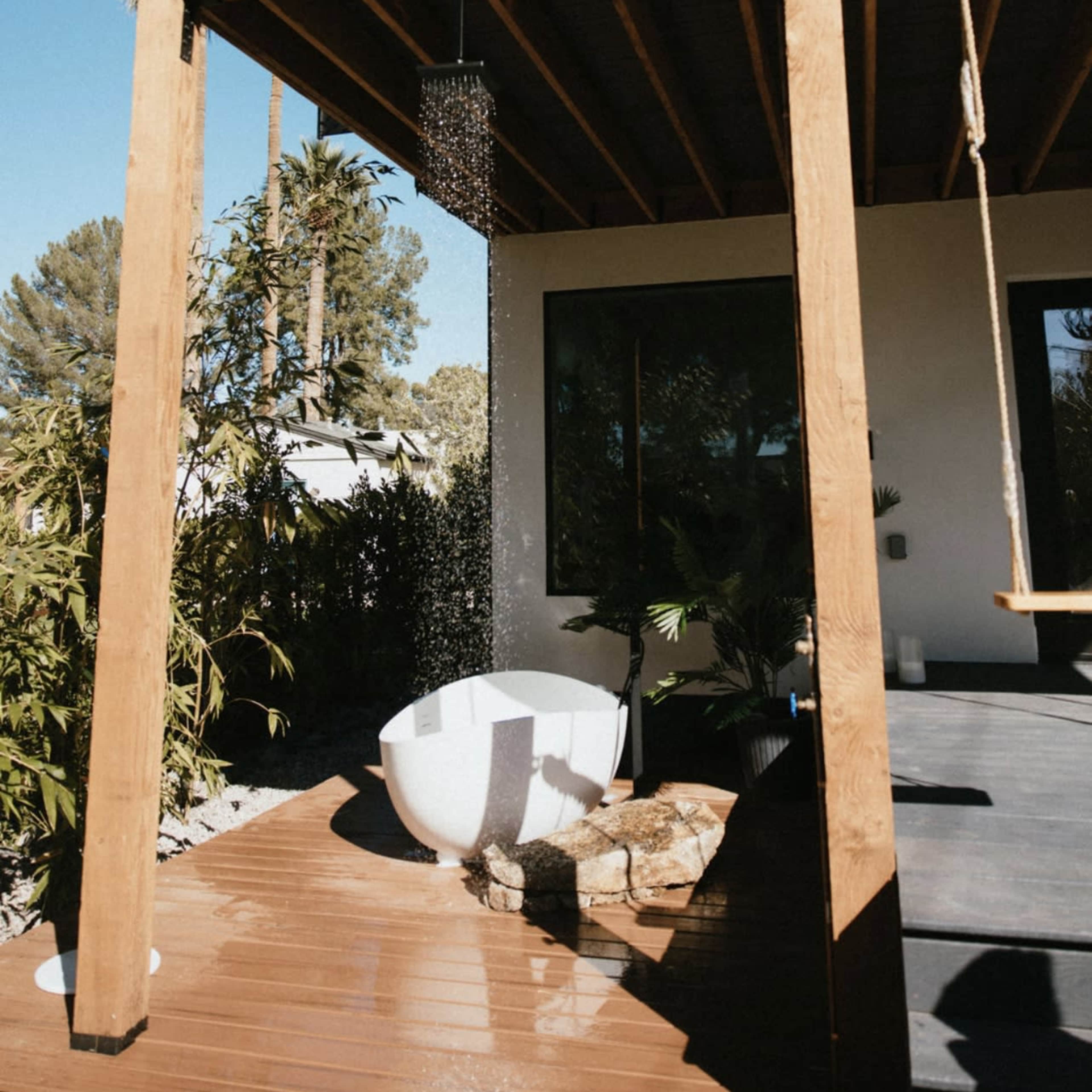 A modern outdoor bathtub is situated beneath a wooden deck, surrounded by greenery and features a rain shower overhead.