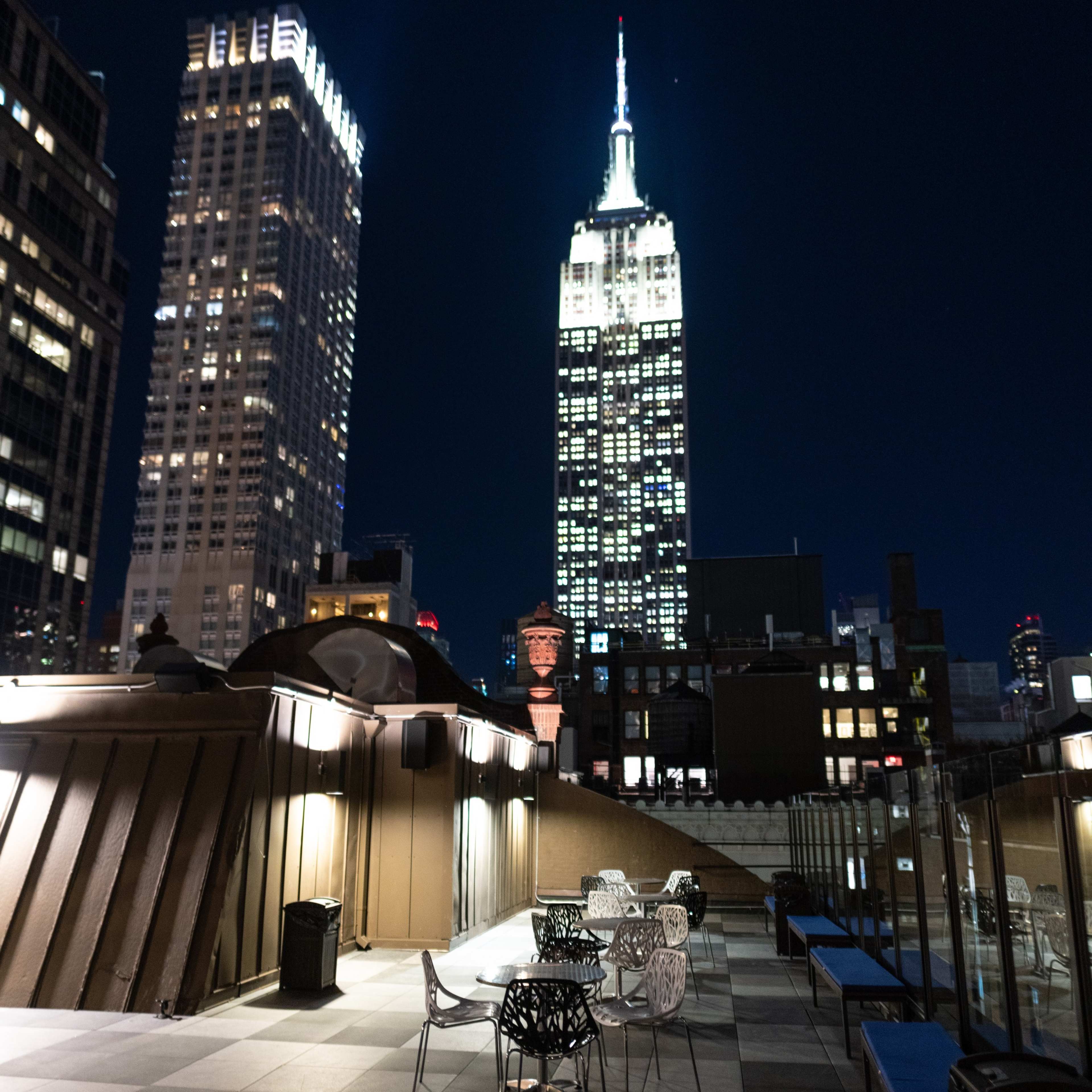 A rooftop terrace with chairs and tables, overlooking the illuminated Empire State Building and surrounding skyscrapers at night.