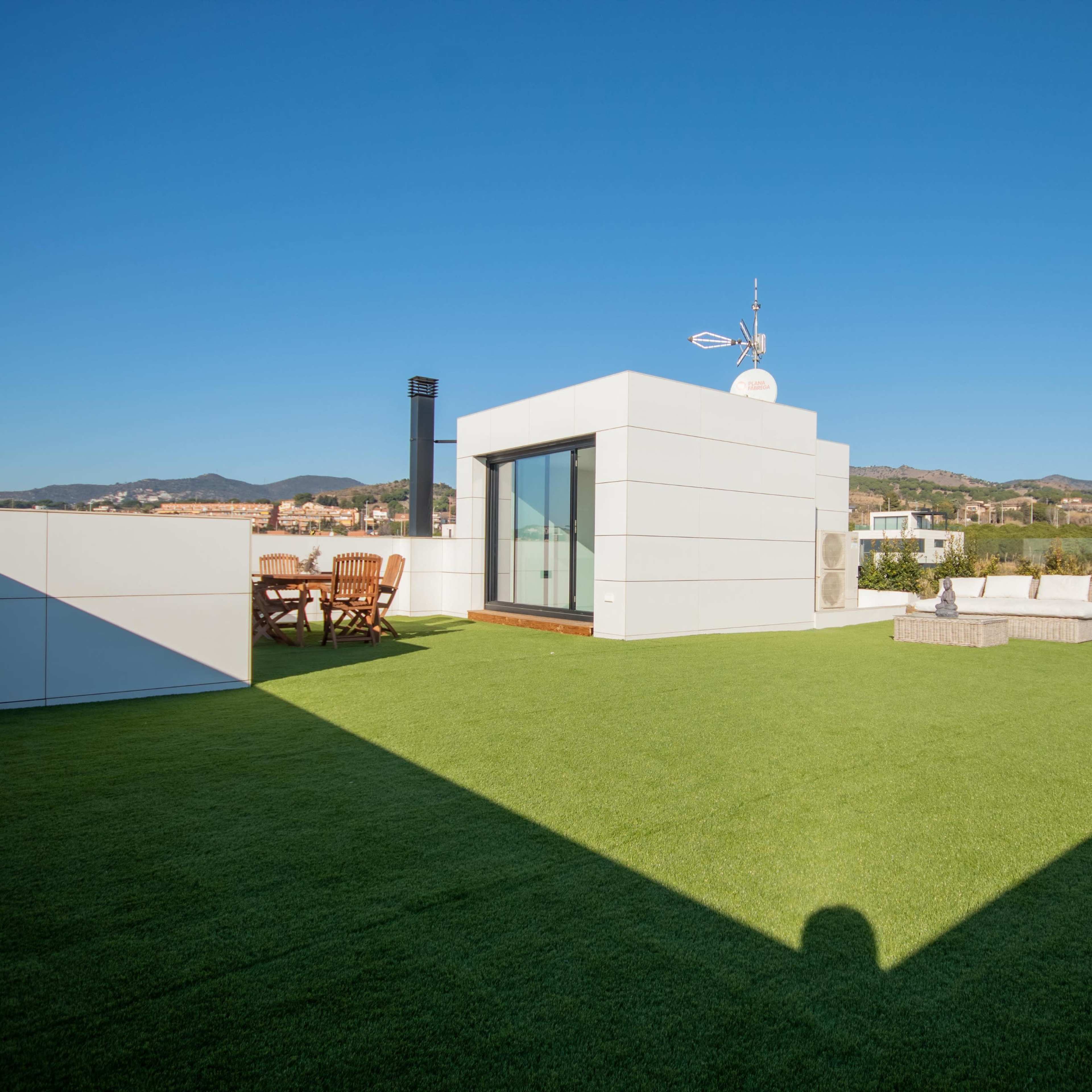 A rooftop terrace with green artificial grass, modern furniture, and a clear blue sky in the background.