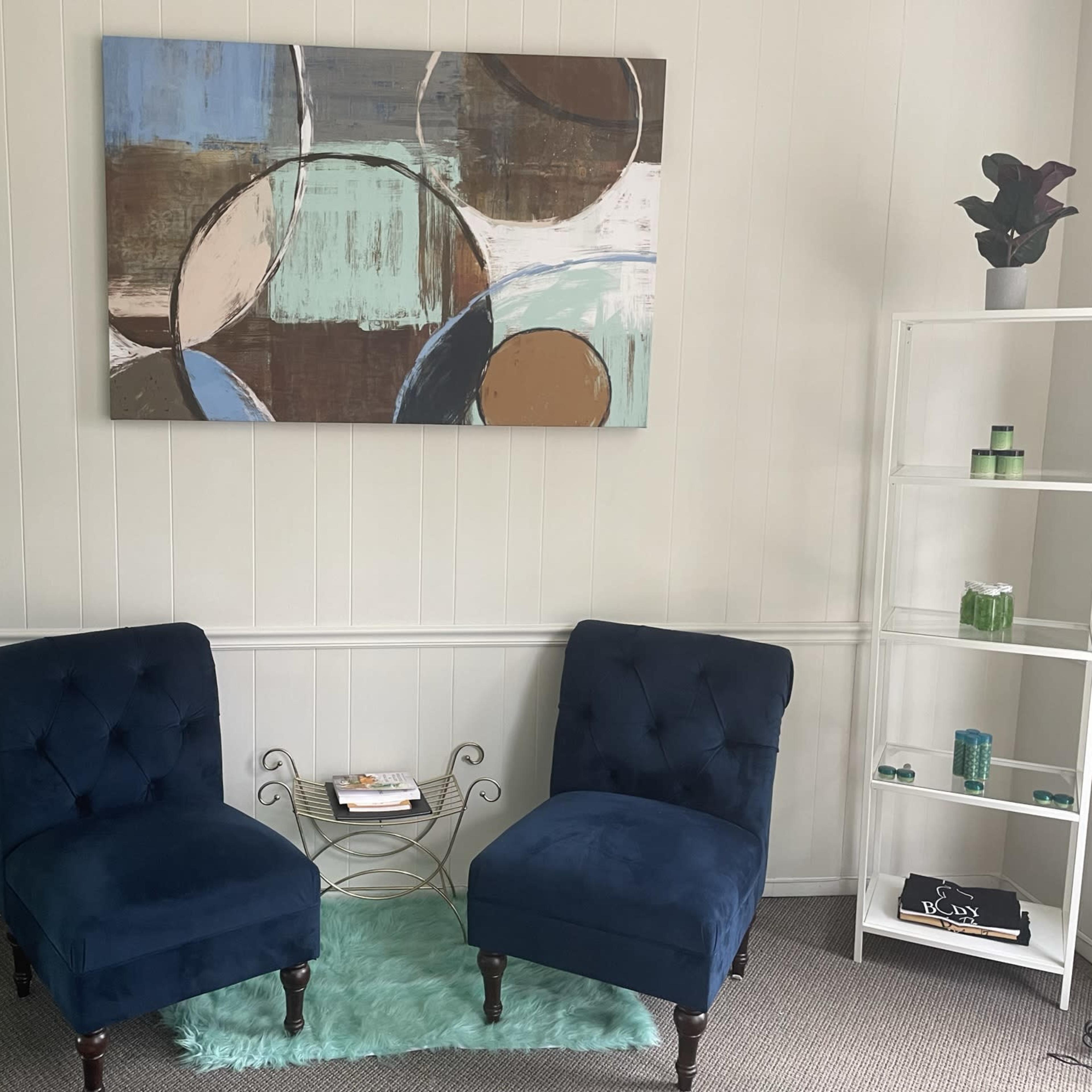 A small, neatly arranged seating area with two blue chairs, a circular table, and shelves displaying decorative items and plants.