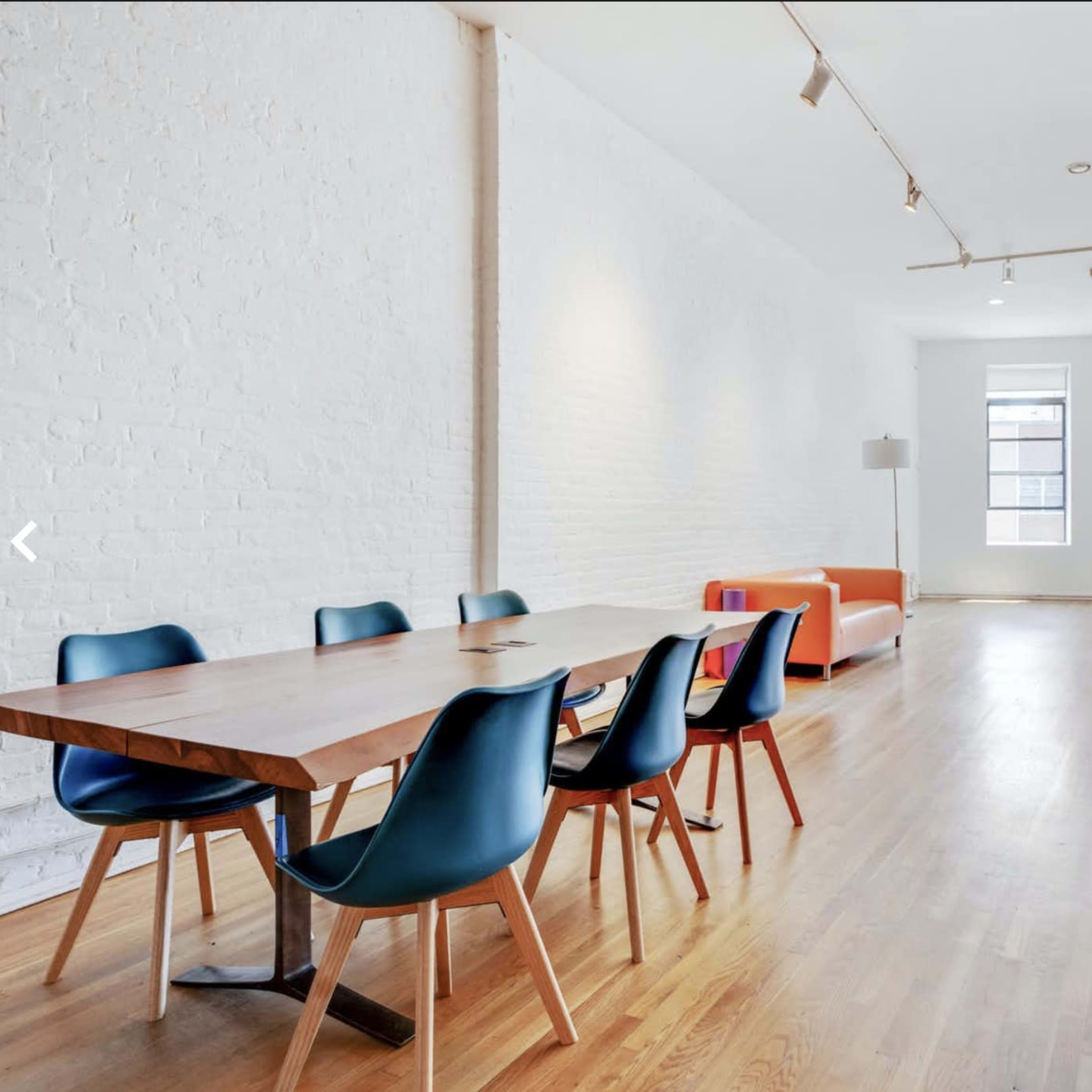 A spacious room with a wooden dining table, six chairs, and an orange couch against a white brick wall.
