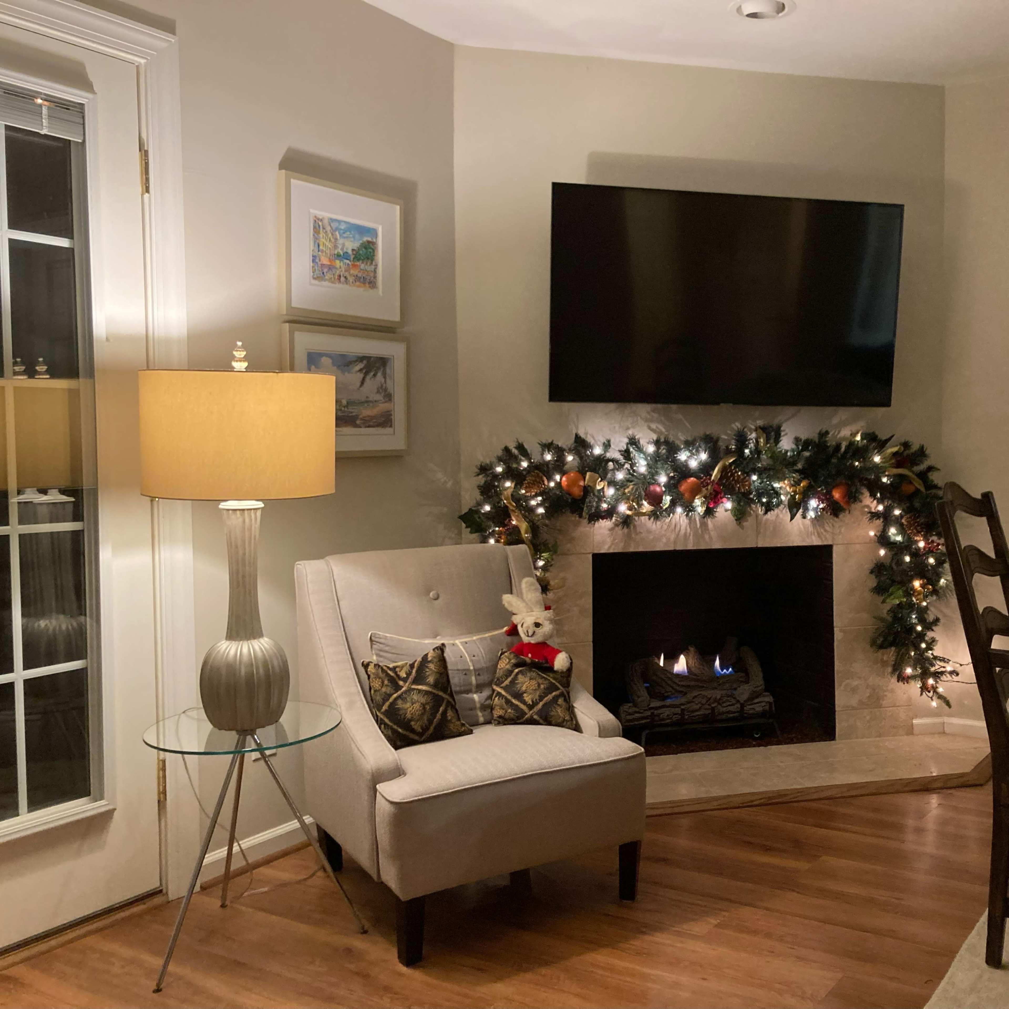 A cozy living room corner with a fireplace adorned with garland, a television mounted on the wall, and a chair beside a side table and a lamp.