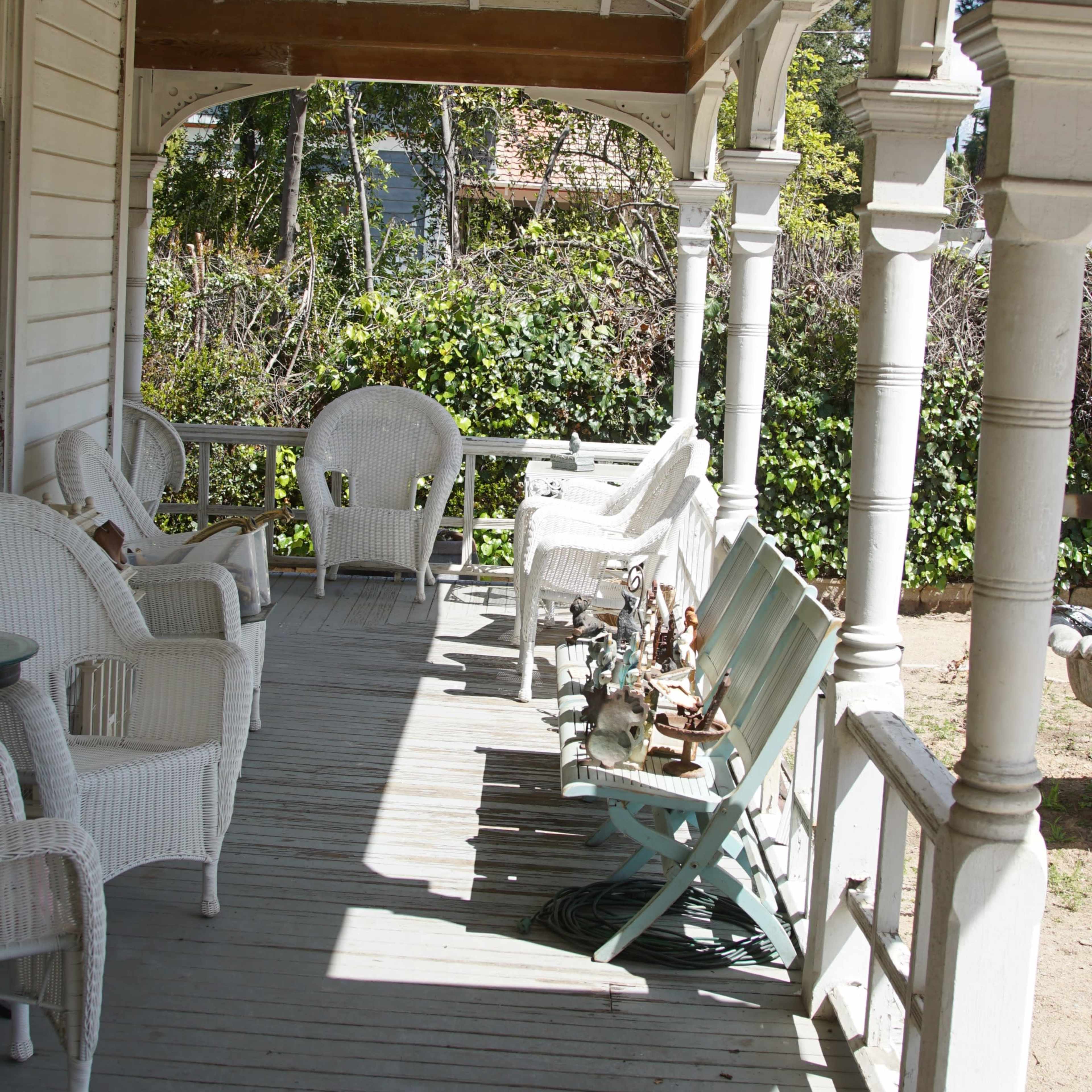 A covered porch with white wicker chairs and wooden flooring, surrounded by greenery.