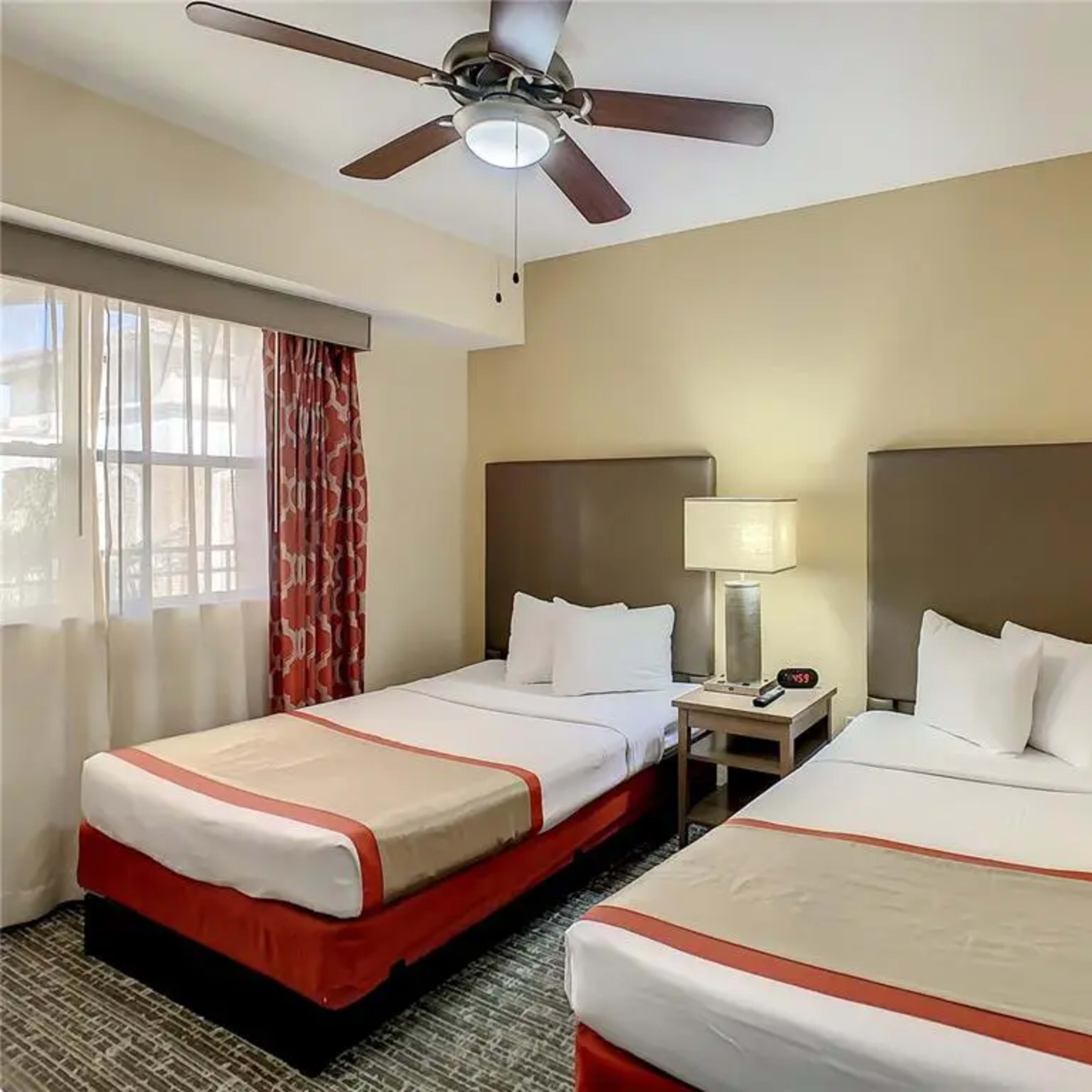 A hotel room with two beds, a bedside table, a lamp, and a ceiling fan, illuminated by natural light from a window.