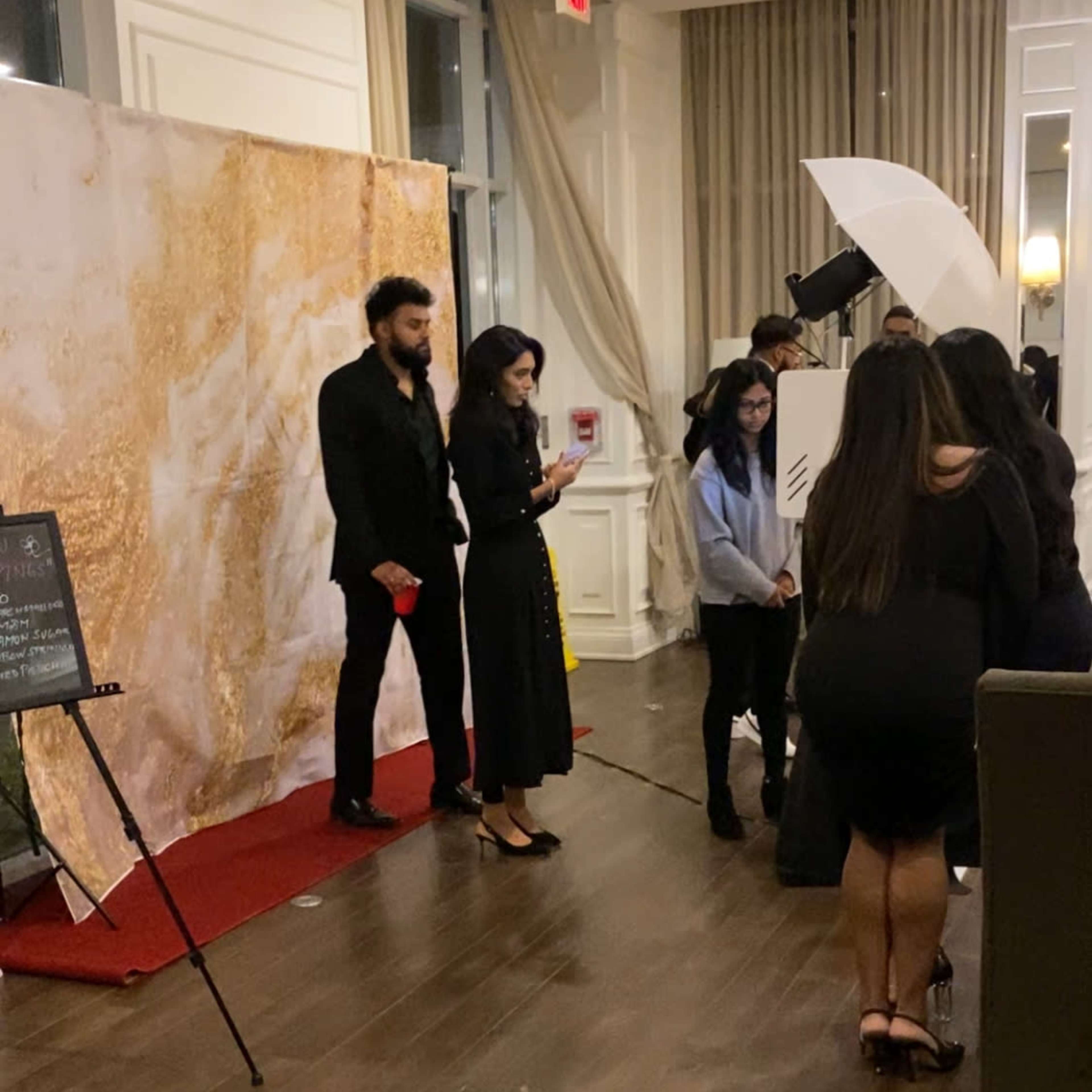 A group of people is gathered in an elegantly decorated indoor space with a gold and white backdrop, while two individuals stand by a red carpet.