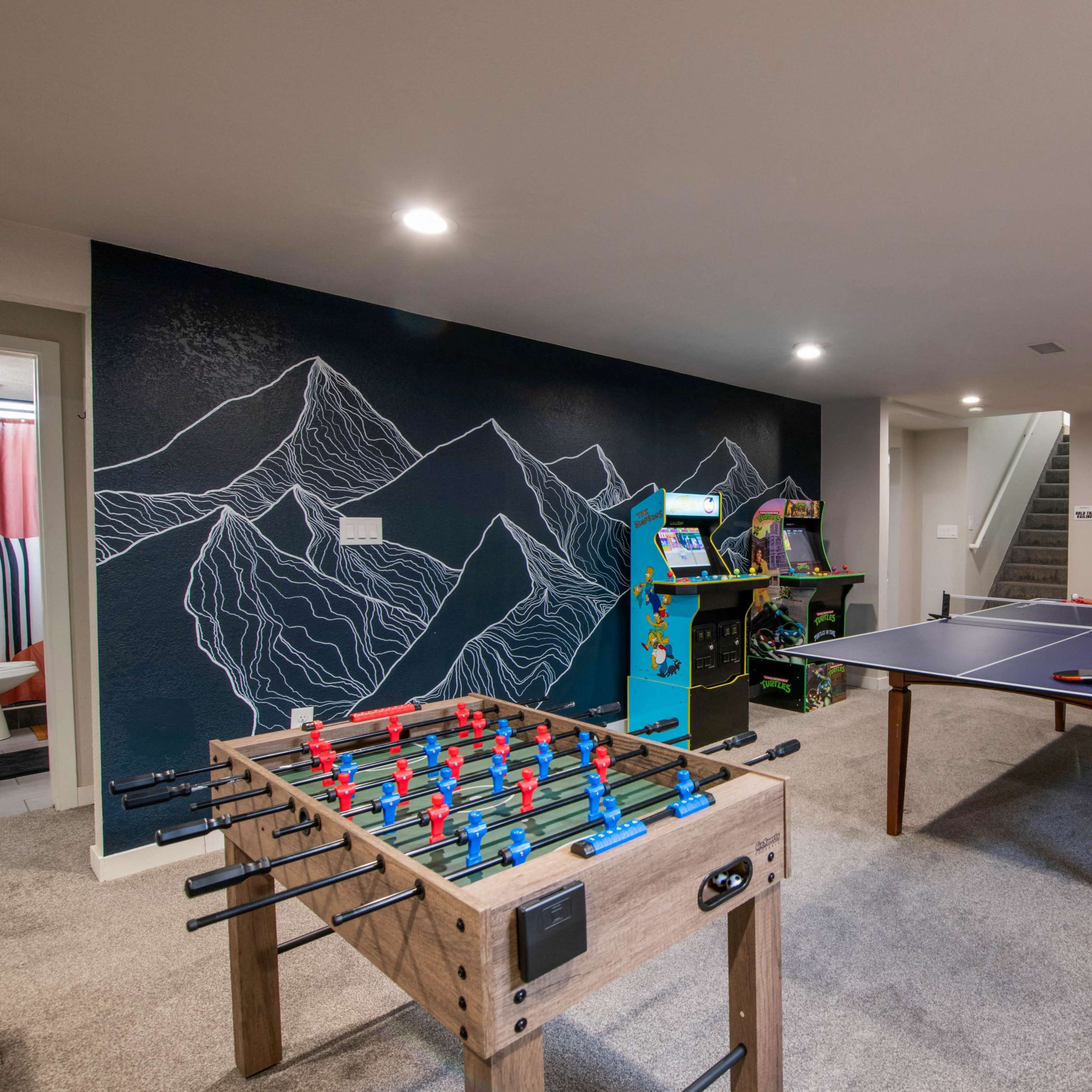 A basement game room featuring a foosball table, an arcade machine, a ping pong table, and a mountain mural on the wall.