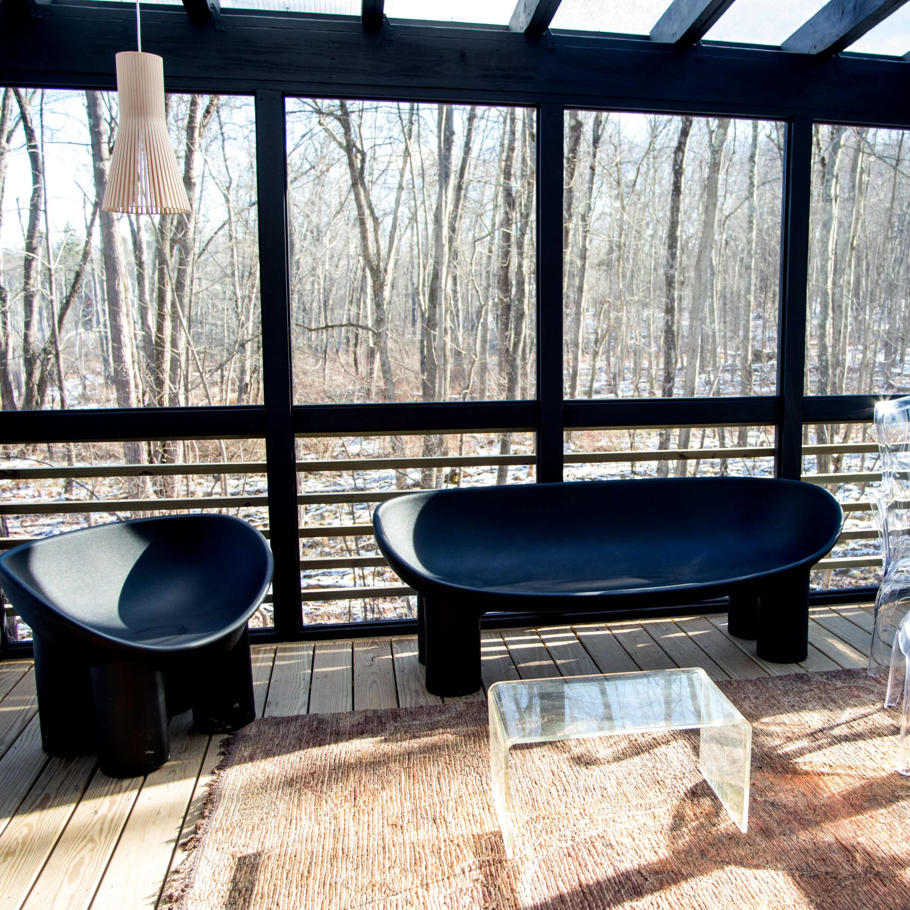A screened porch with two black modern chairs, a clear acrylic table, and large windows overlooking a wooded area.