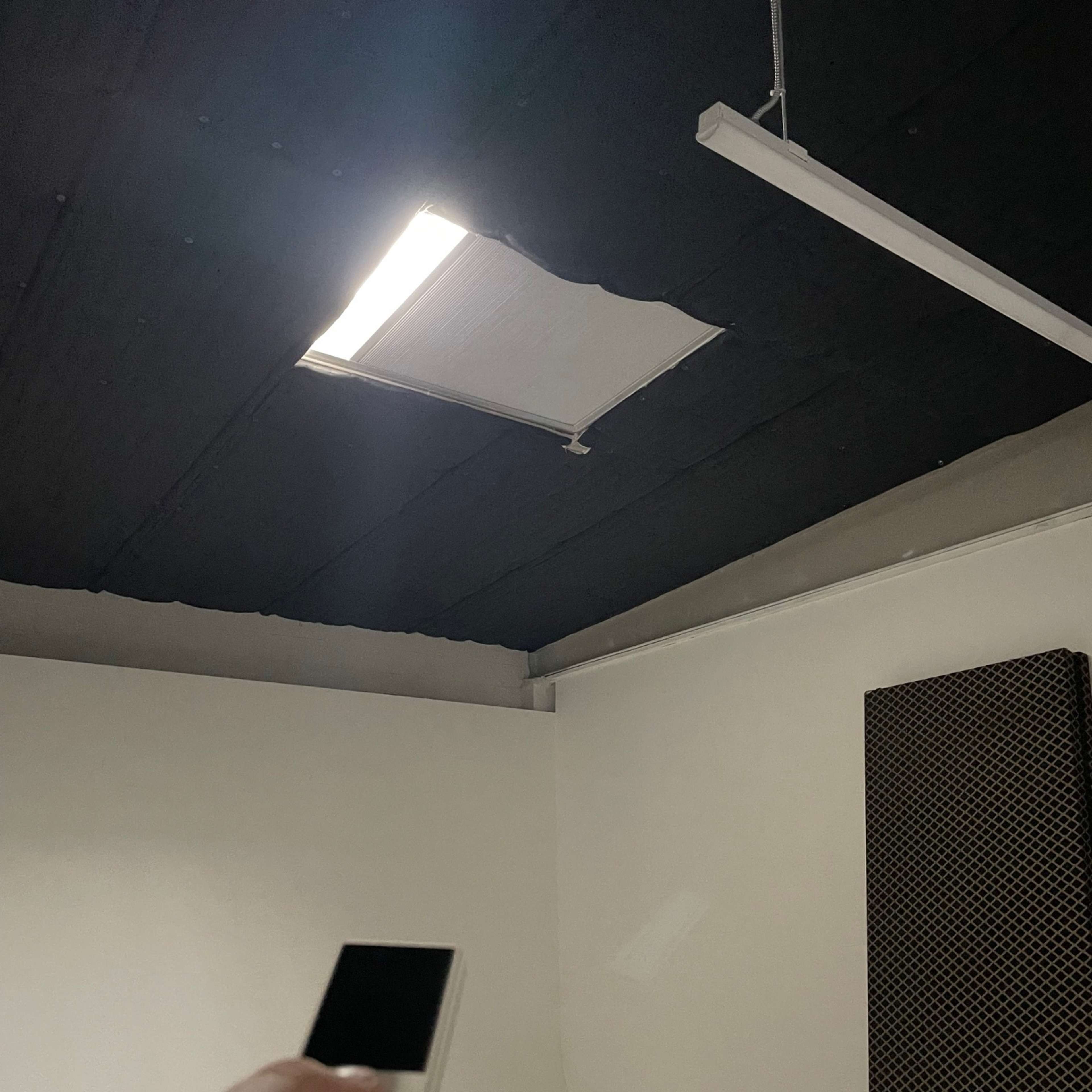 A hand holds a remote control aimed at a ceiling light fixture above an open section of the ceiling.