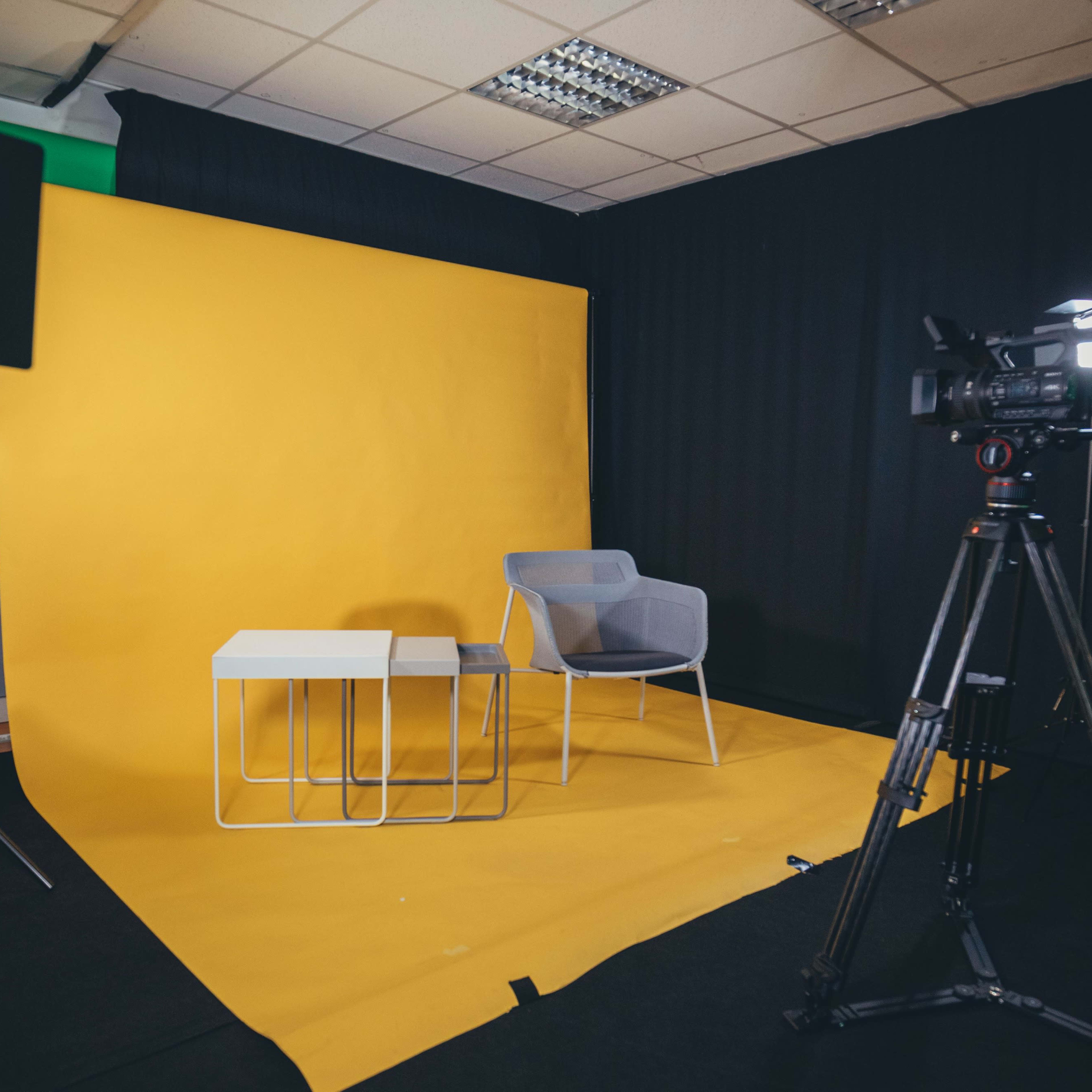 A photography or video studio setup with a yellow backdrop, a gray chair, two white tables, and equipment for recording on the right.