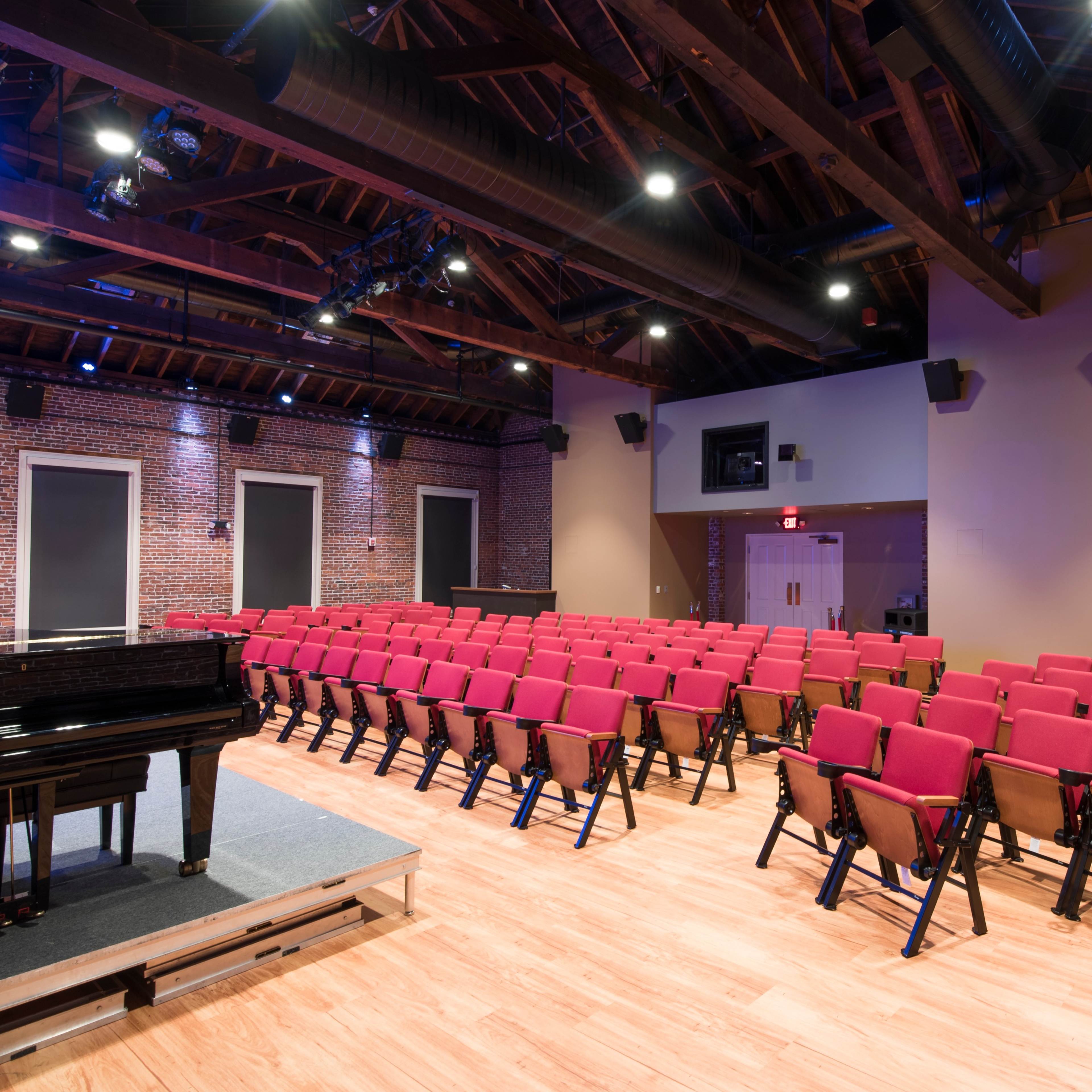 A concert hall with red seats arranged in rows facing a black grand piano on a stage, illuminated by overhead lights.