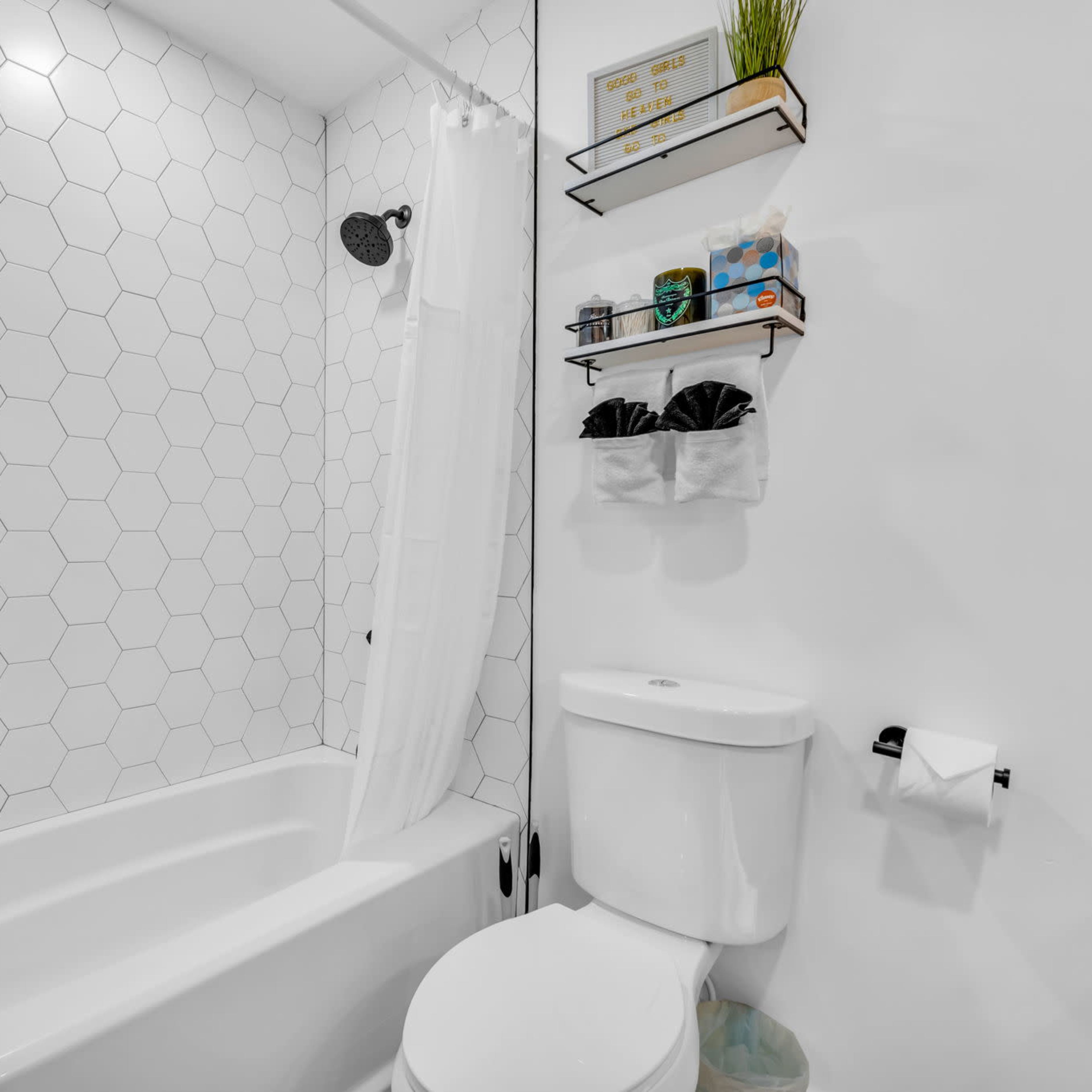A modern bathroom featuring a white bathtub with a showerhead, hexagonal tile walls, a toilet, and shelves holding toiletries.