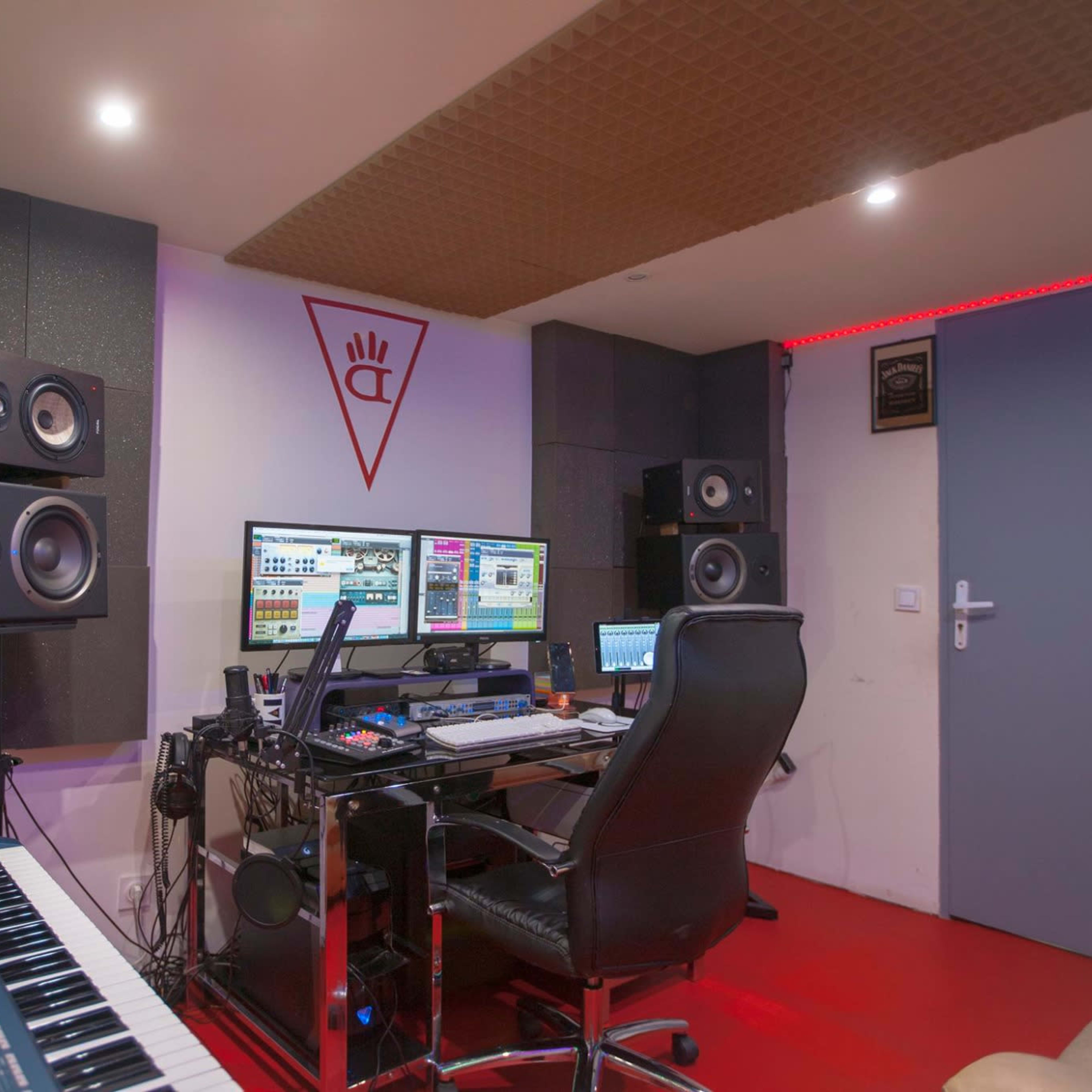 A music production studio featuring a desk with two computer monitors, studio speakers, a keyboard, and soundproofing on the walls.
