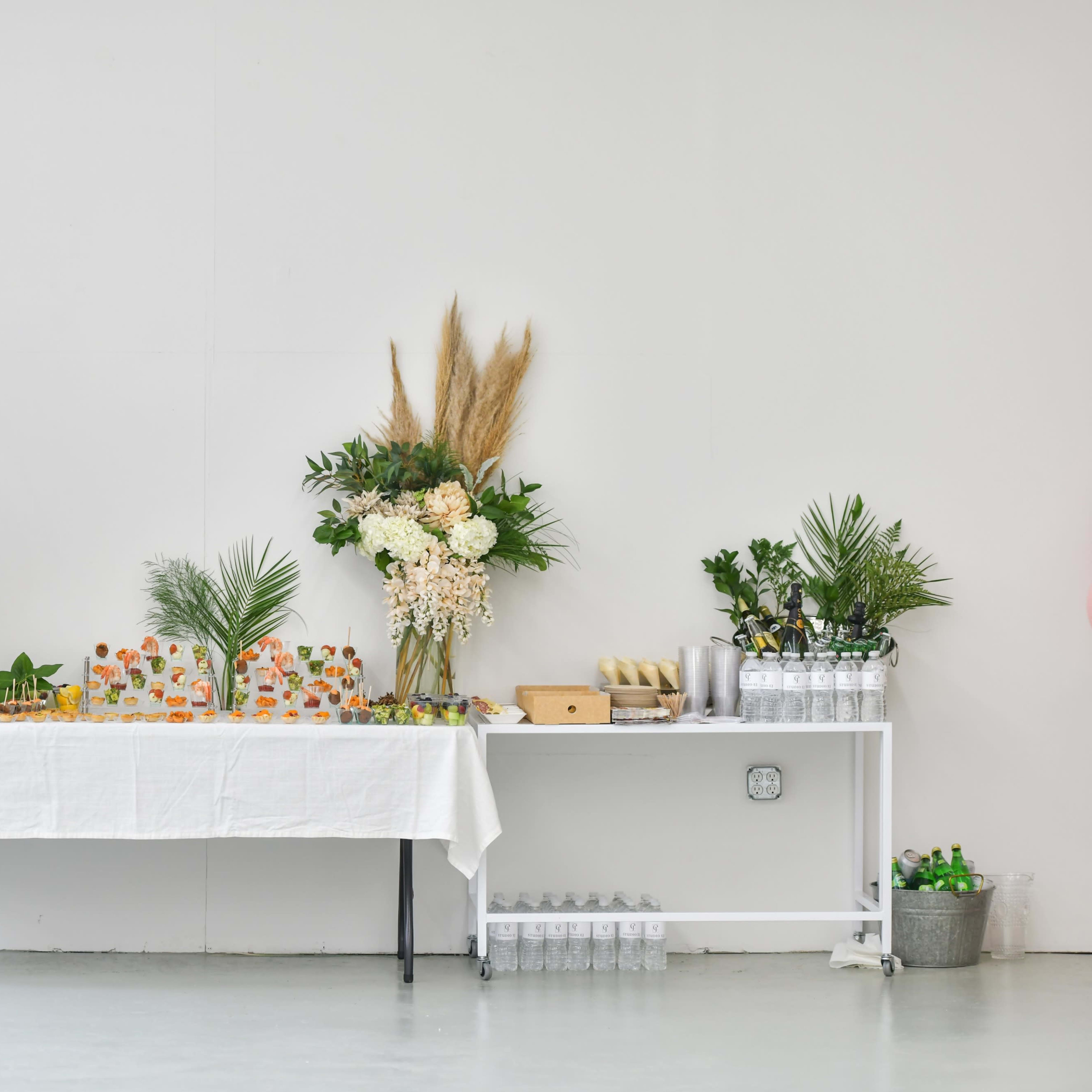 A table with a white cloth is set up against a white wall, displaying an assortment of drinks and snacks, while potted plants add greenery to the scene.