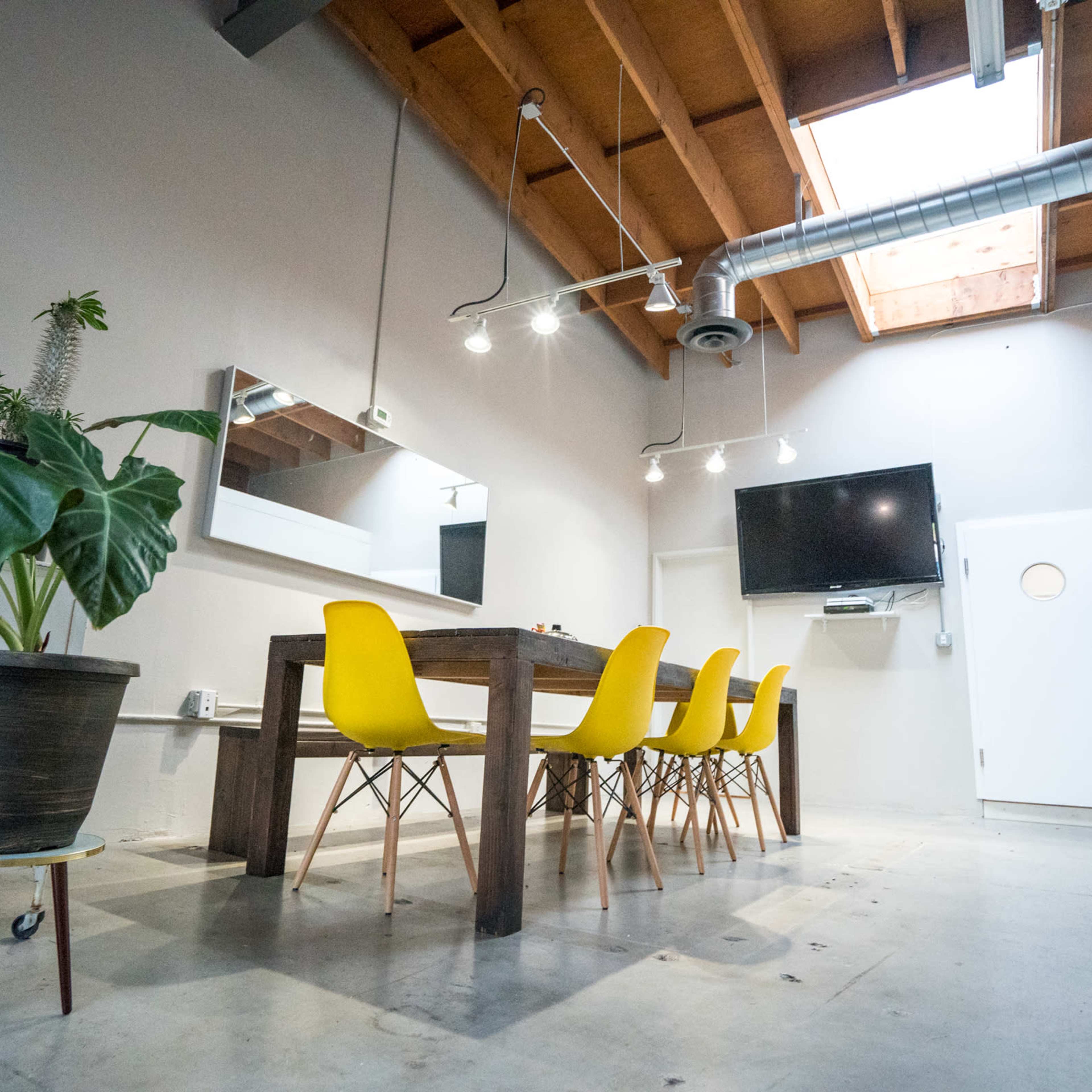 A modern conference room features a long wooden table surrounded by yellow chairs, large mirror, wall-mounted TV, and a skylight providing natural light.
