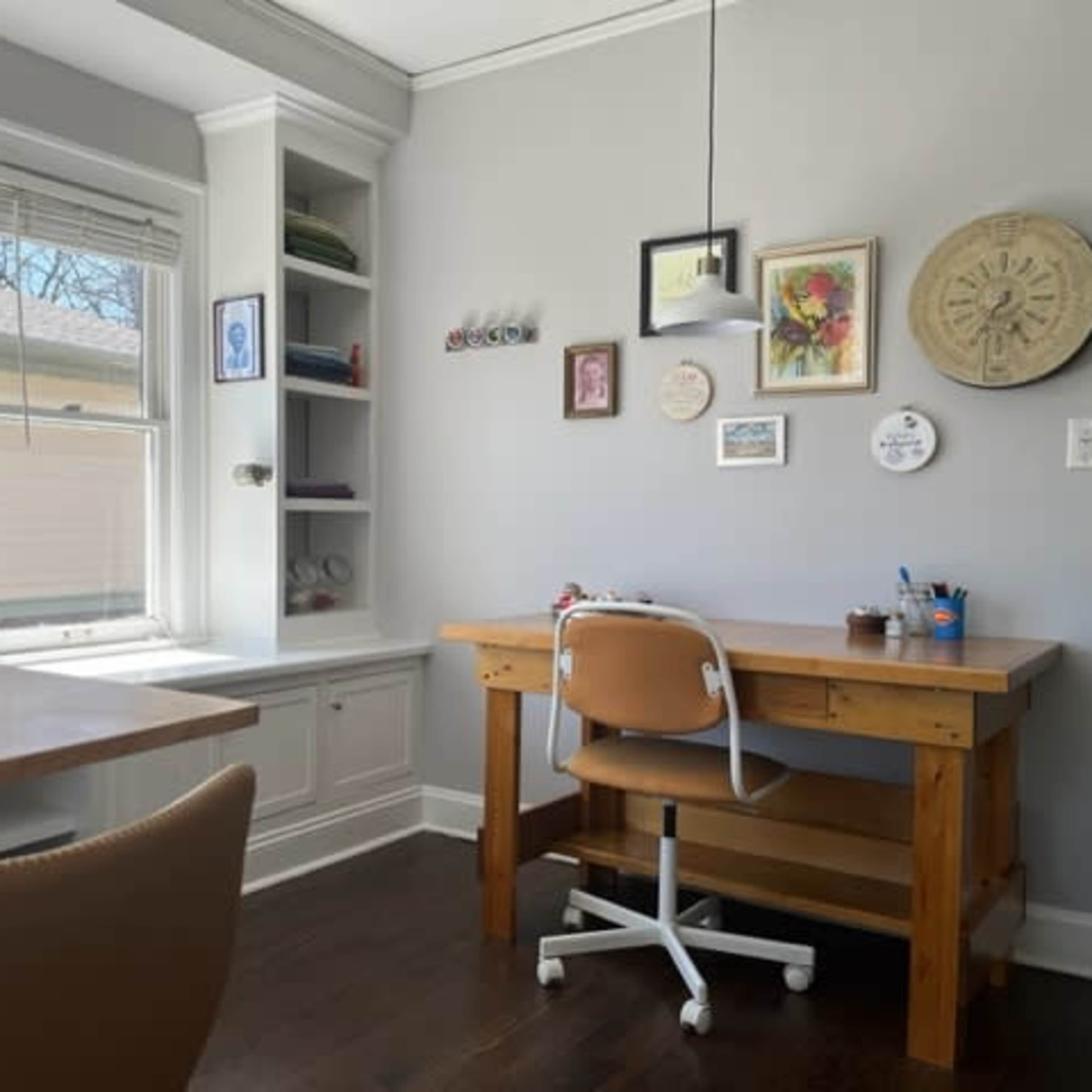 A bright room with two tables, one featuring a wooden desk and chair, and a shelf with framed pictures and decorative items against a light gray wall.