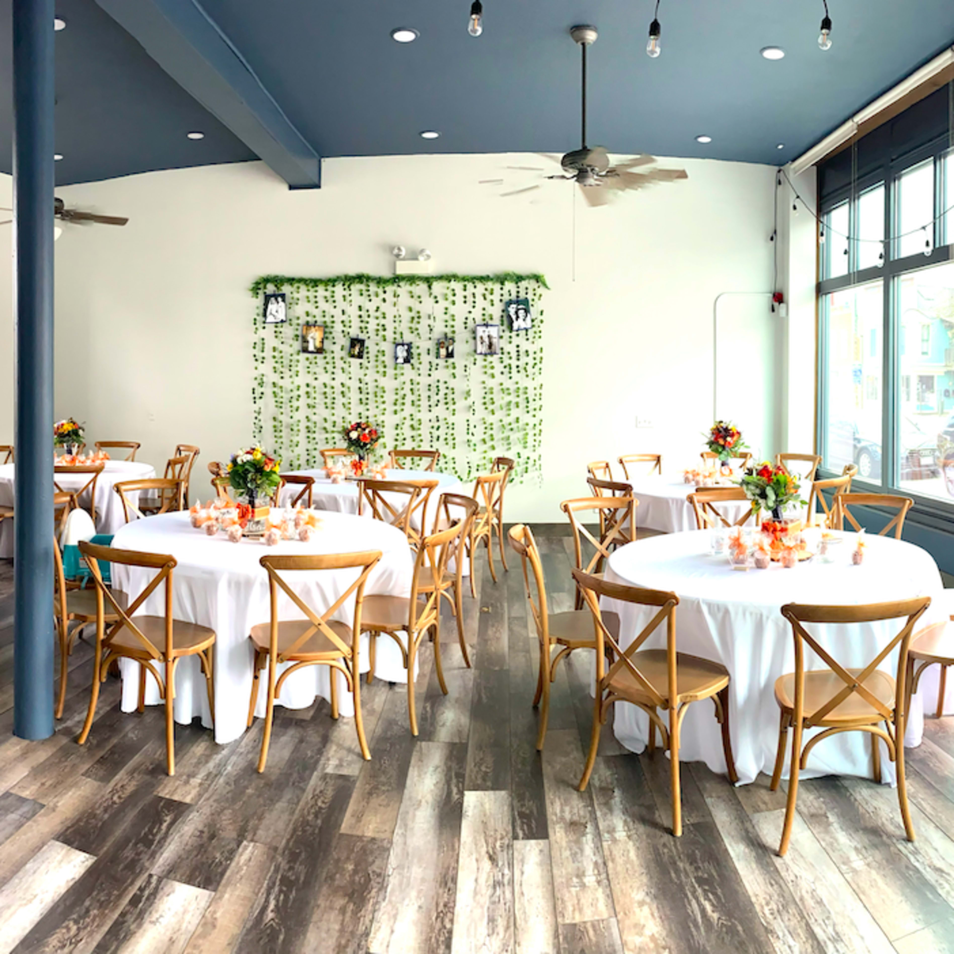 A decorated event space with round tables, white tablecloths, and centerpieces, surrounded by large windows and a greenery backdrop.