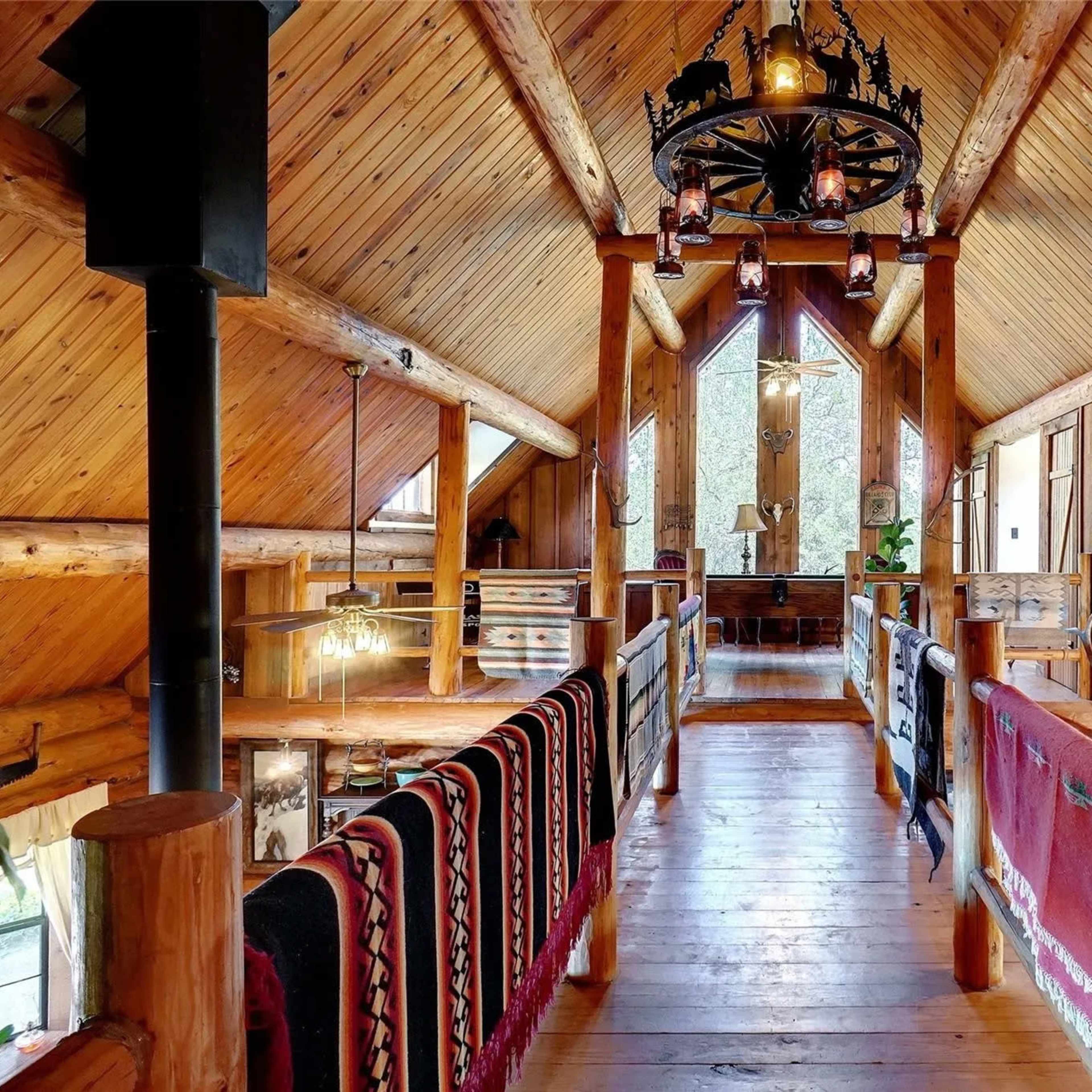 A rustic wooden loft with exposed beams, a high ceiling, and a balcony overlooking a spacious area below, adorned with decorative items and rugs.