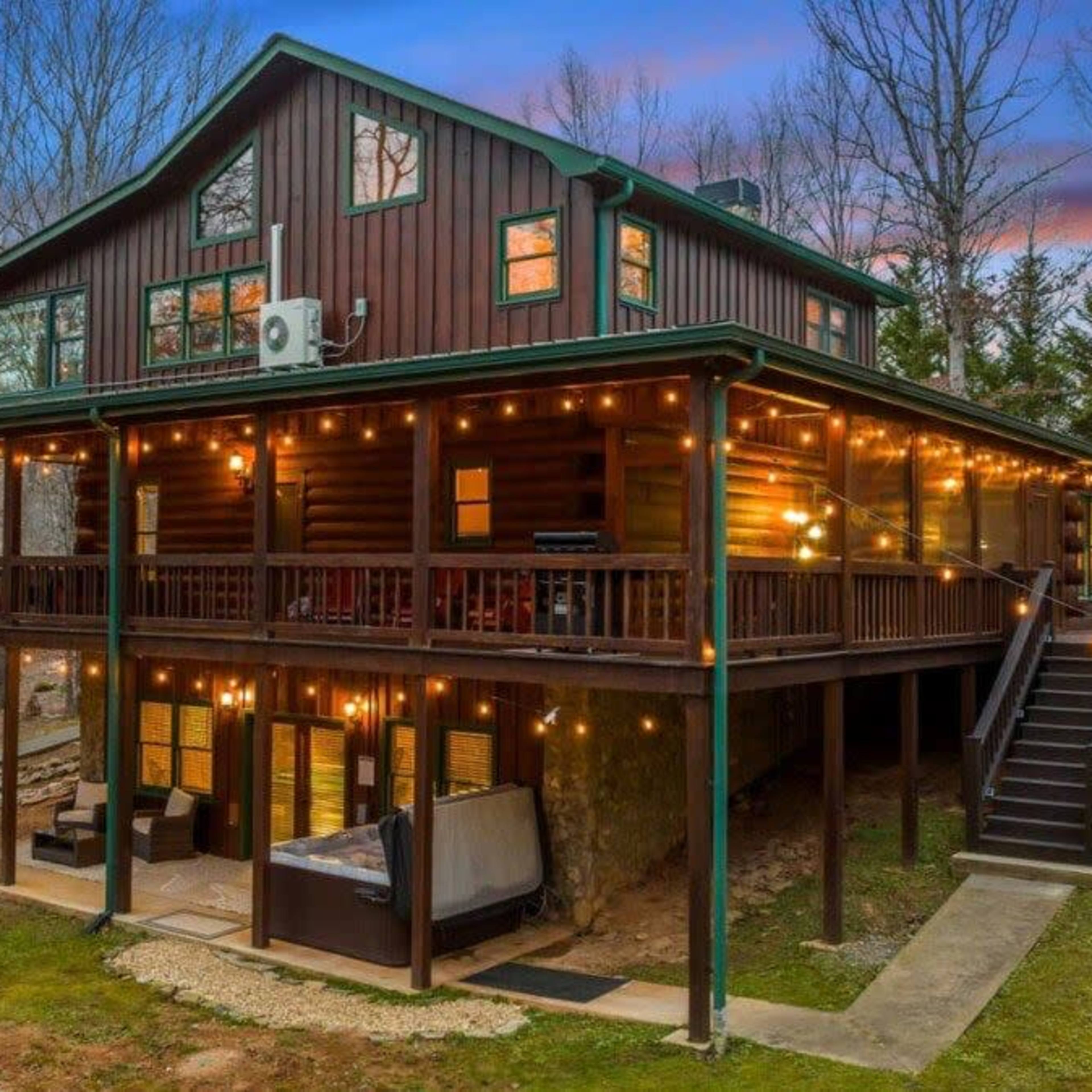 A large wooden cabin with multiple levels and string lights is surrounded by trees in a wooded area.