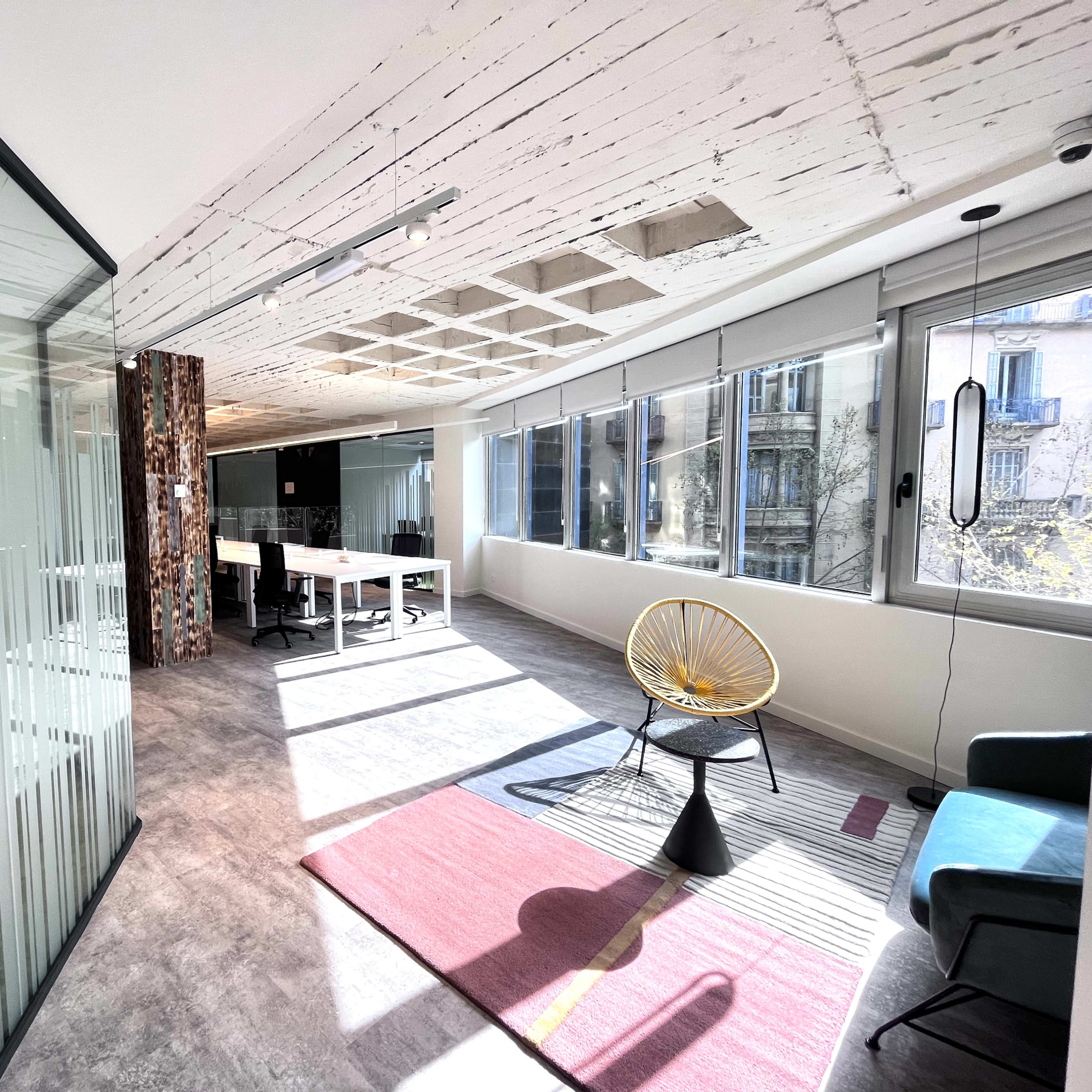 A modern office space with large windows, a glass partition, and a mix of seating options including a chair and a table.