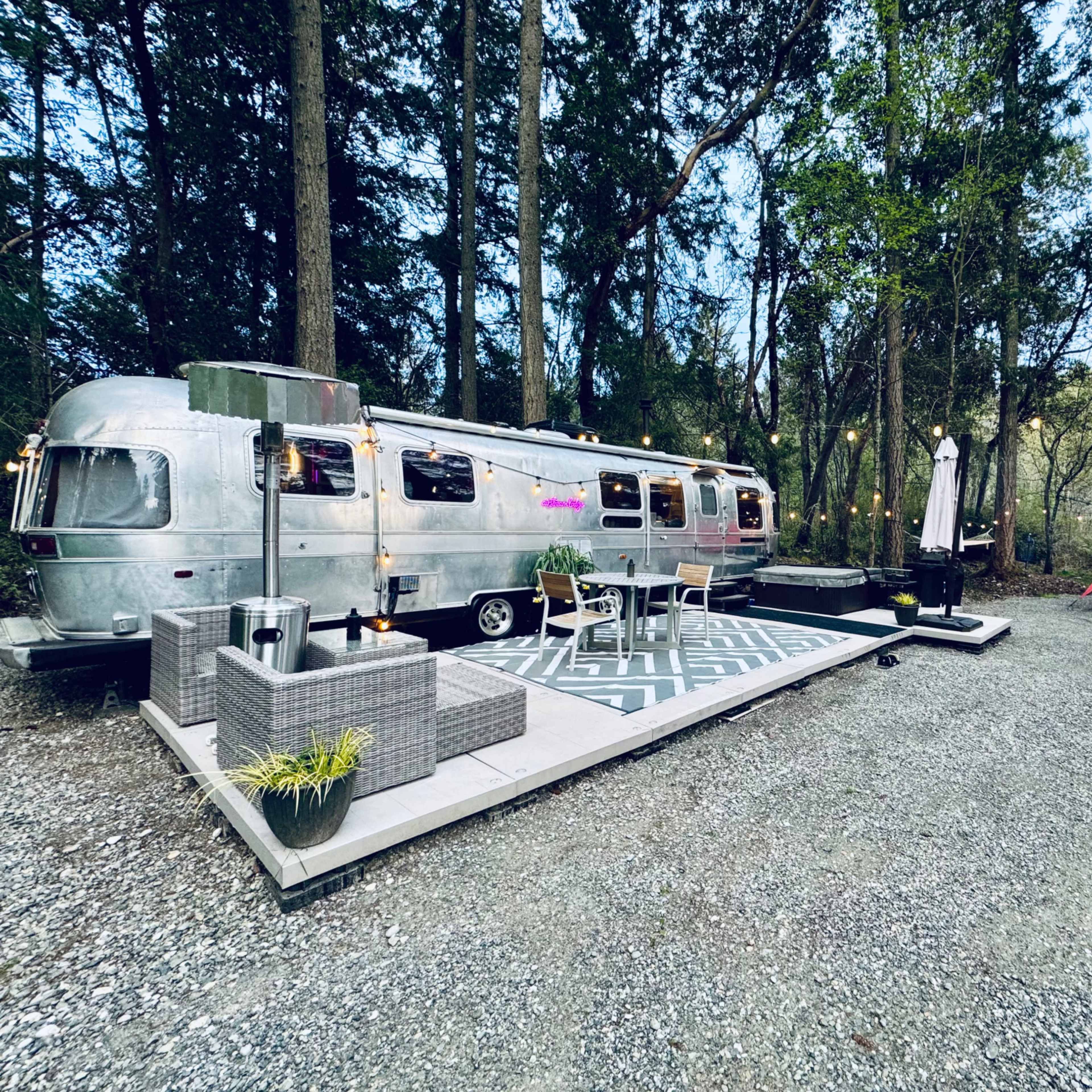 A vintage Airstream trailer is parked on a gravel area surrounded by trees, with an outdoor seating arrangement and a red canopy nearby.