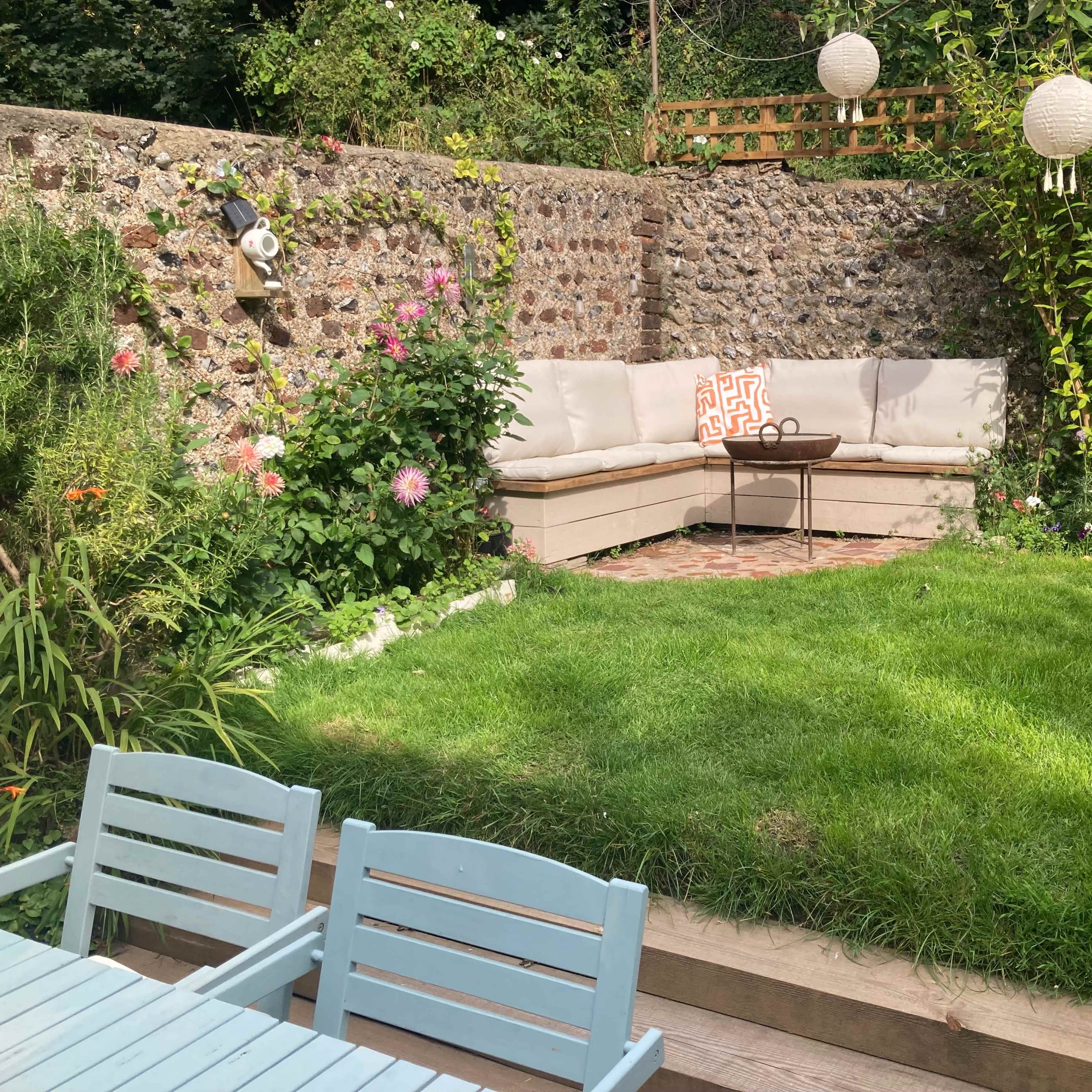 A cozy seating area with a sectional couch and a small table is nestled in a landscaped garden surrounded by a stone wall and floral plants.