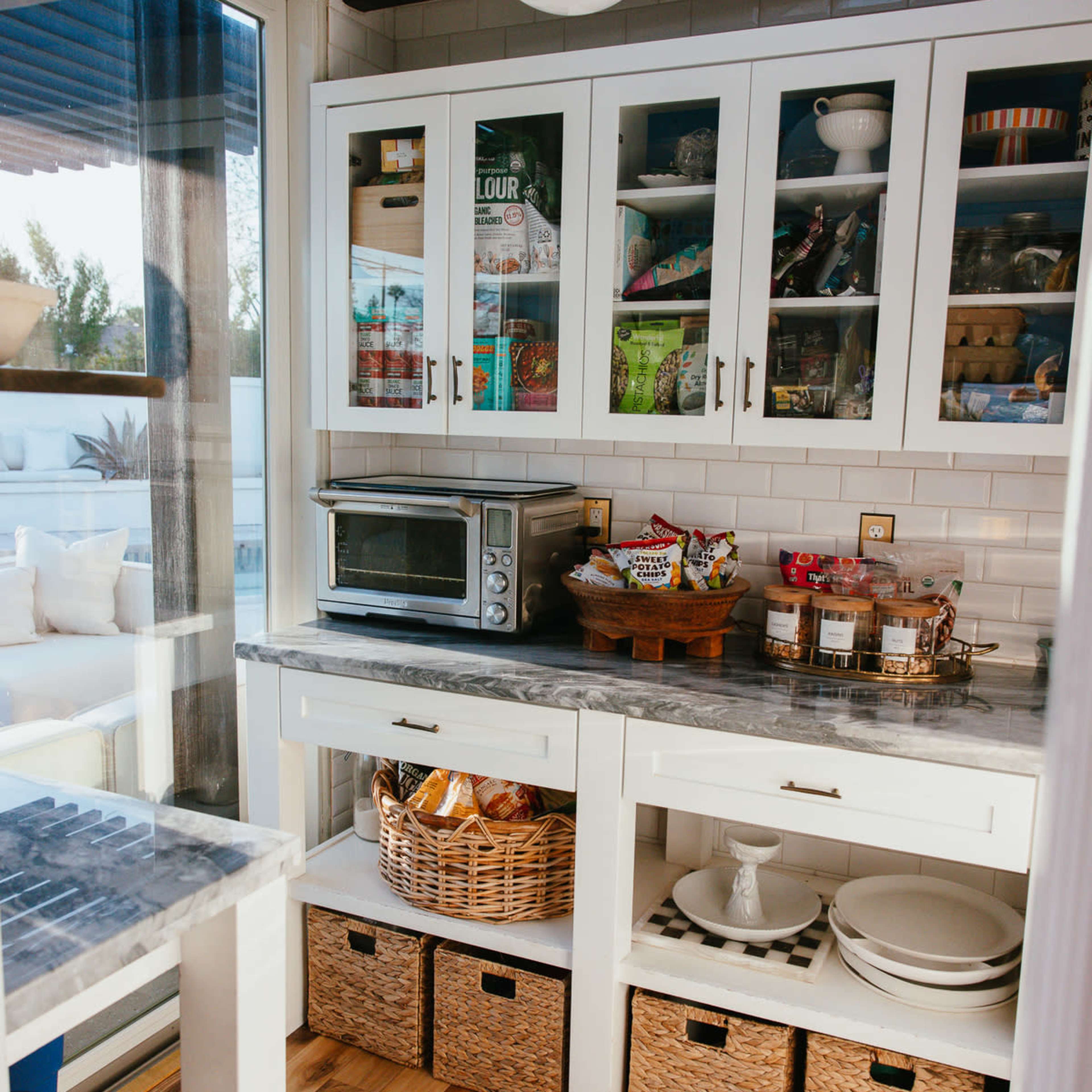 The kitchen features white cabinets with glass fronts, showcasing various dishes and jars, while a marble countertop sits above woven storage baskets.